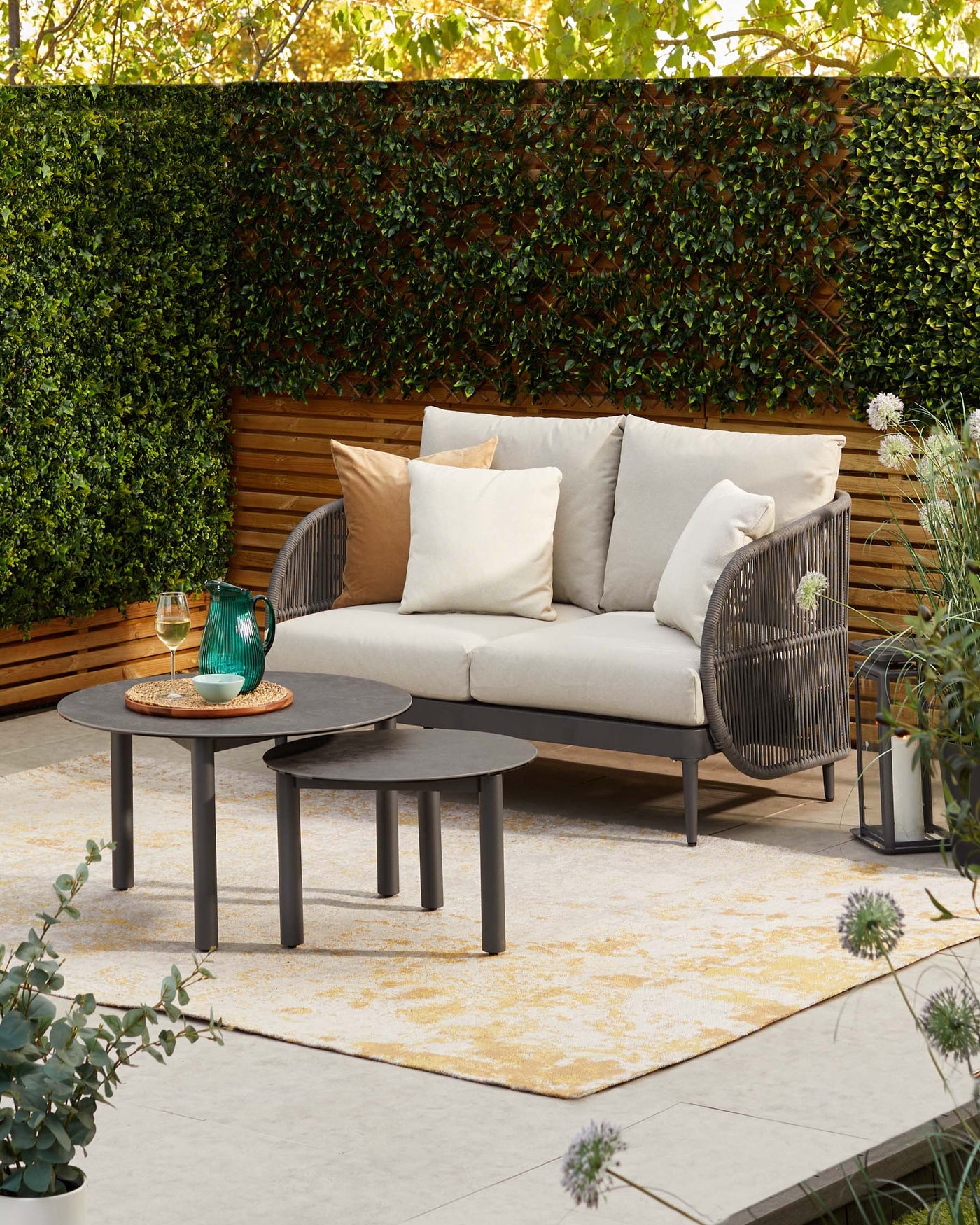 Elegant outdoor patio setup featuring a modern two-seater sofa with beige cushions and wicker side panels, accompanied by two round nesting coffee tables with dark matte finishes, on a textured cream and yellow area rug.