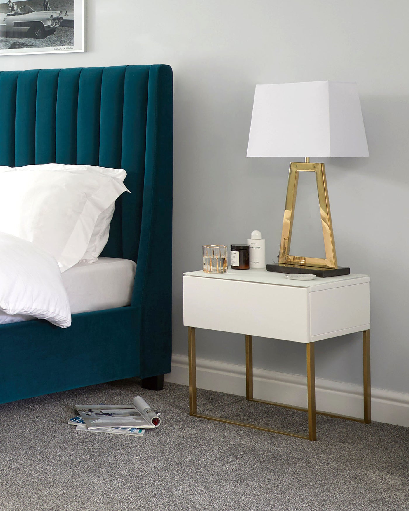 Contemporary bedroom furniture featuring a teal upholstered tall headboard with vertical channel tufting, and a modern white bedside table with a smooth finish and brass legs. The table is adorned with a geometric brass table lamp with a white shade, alongside a neatly arranged selection of cosmetics and books.