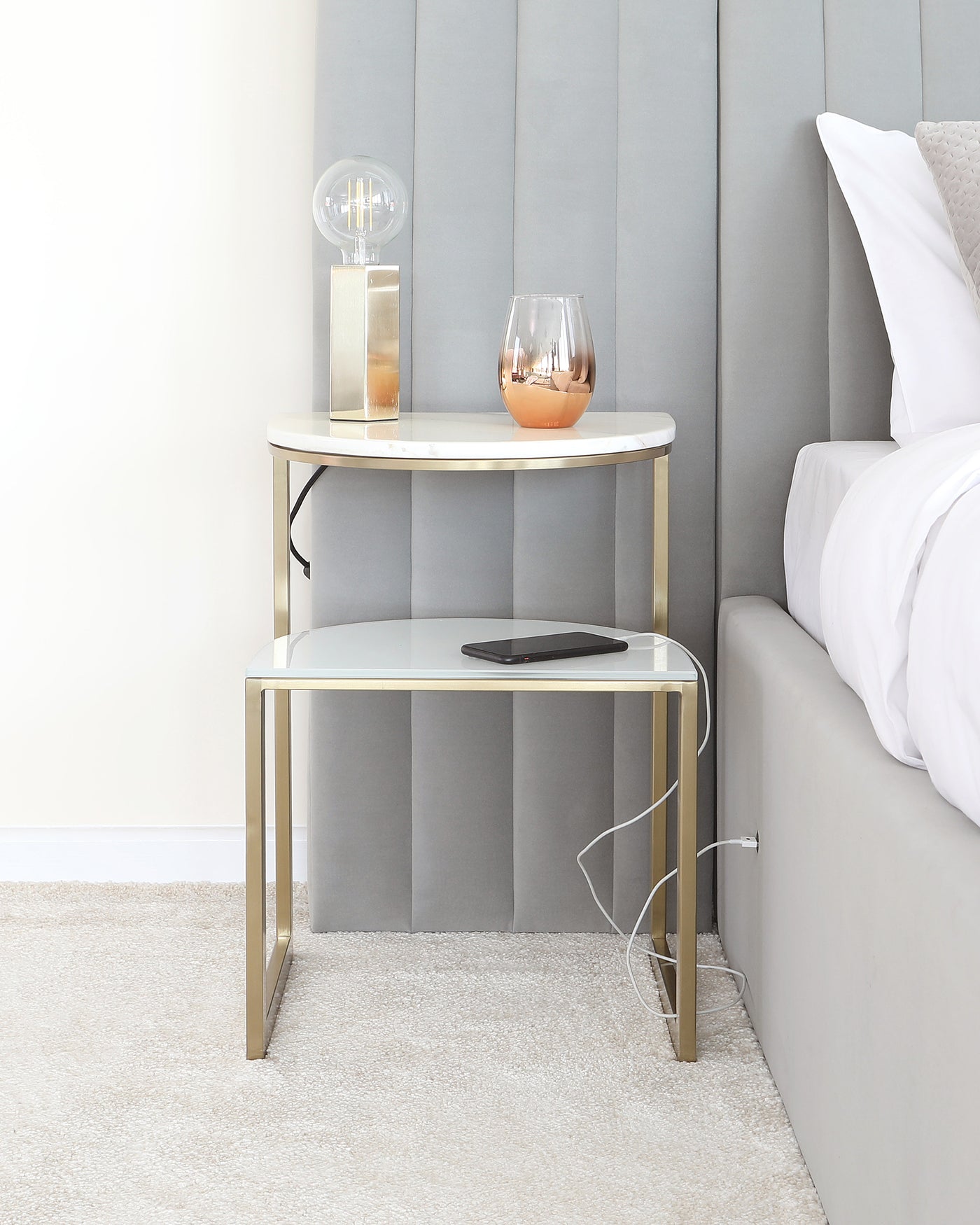 A modern two-tiered round side table with a white marble top and gold-finished metal frame, positioned next to a bed with grey padded headboard on a textured beige carpet. The upper tier supports a decorative lamp and a stemless wine glass, while the lower tier holds a smartphone with a charging cable.