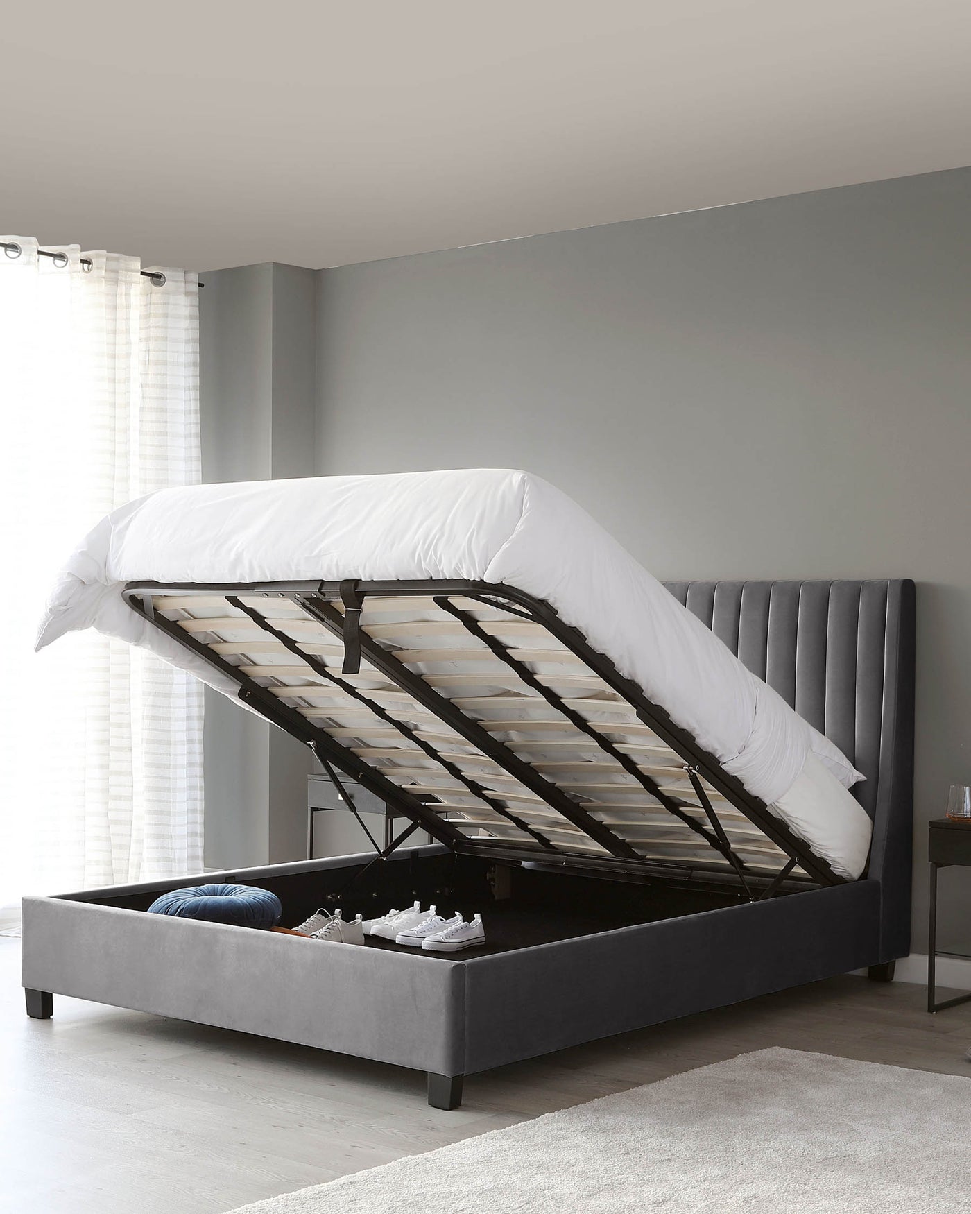 A contemporary-style grey upholstered storage bed with a lifting frame to reveal an under-bed storage compartment. The bed features a tufted headboard with vertical stitching and a low-profile platform design, supported by dark wooden legs. A mattress is partly lifted to show the wooden slat support system and items stored underneath, including pillows and sneakers.