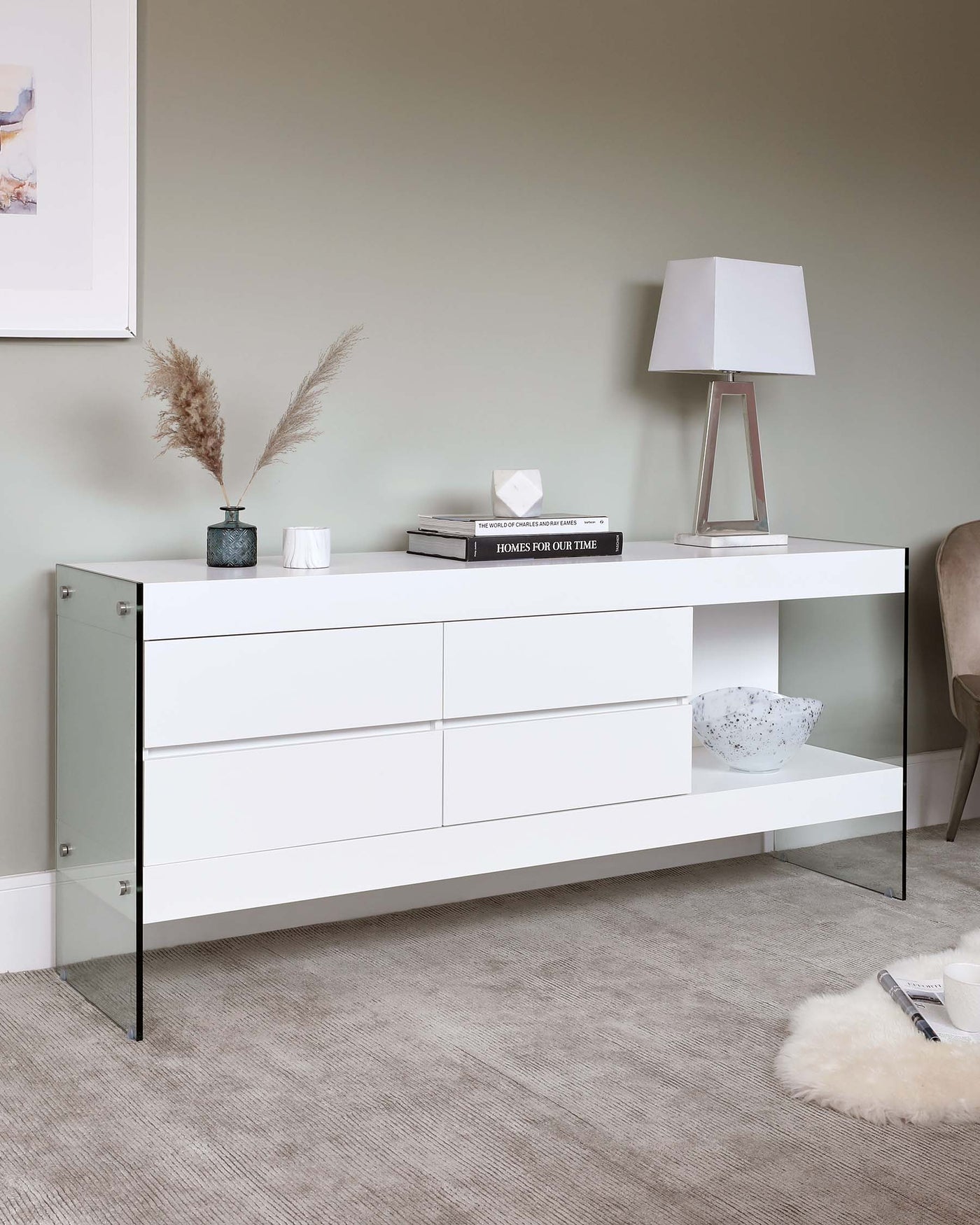 Modern white sideboard with sleek drawers and black metal accents, featuring an integrated shelf space on the right. The piece is styled with a table lamp, decorative books, a vase with dried pampas grass, and a modern textured bowl.