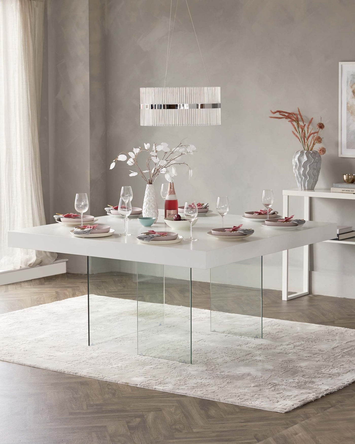 A modern rectangular dining table with a white top supported by four clear glass legs, positioned on a textured white area rug. The table is set with plates, glasses, and cutlery for a meal, and complemented by decorative vases with flowers.