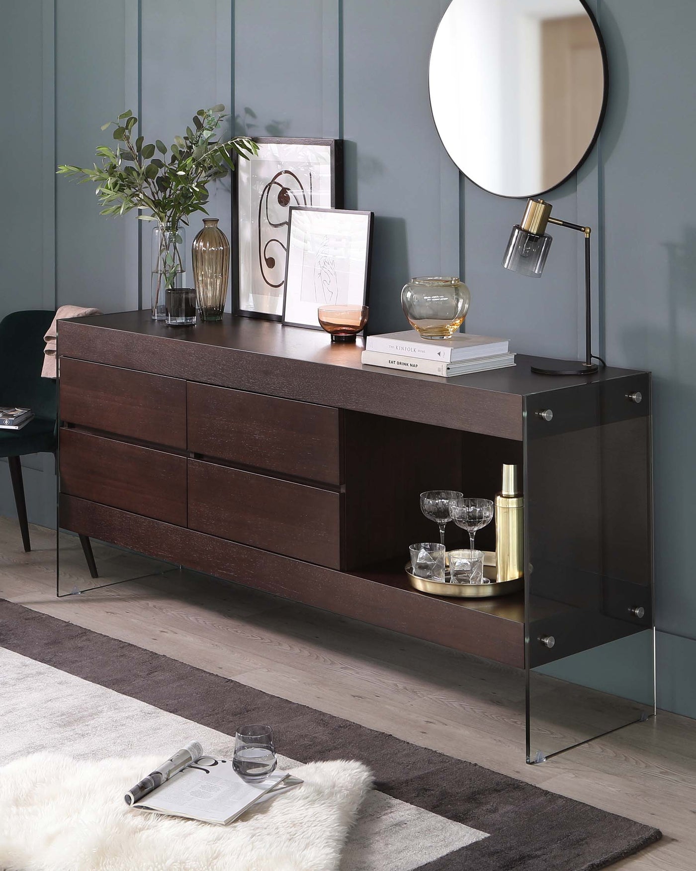 A contemporary wooden sideboard with a rich dark finish, featuring a streamlined silhouette, multiple drawers, and an open shelf with a contrasting gold-tone circular base. The sideboard is supported by a minimalist metal frame with a glass end panel accentuating its modern design.