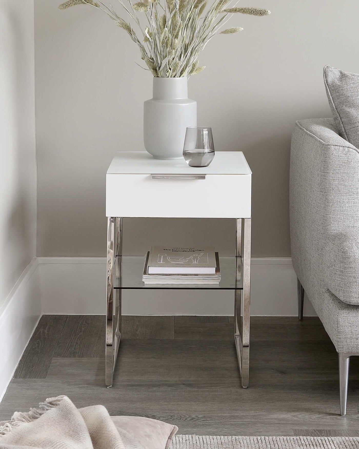 Modern white lacquered side table with chrome legs and an open lower shelf, displayed alongside a grey fabric upholstered armchair. The table is accessorized with a large matte grey vase holding dried foliage and a simple clear glass.