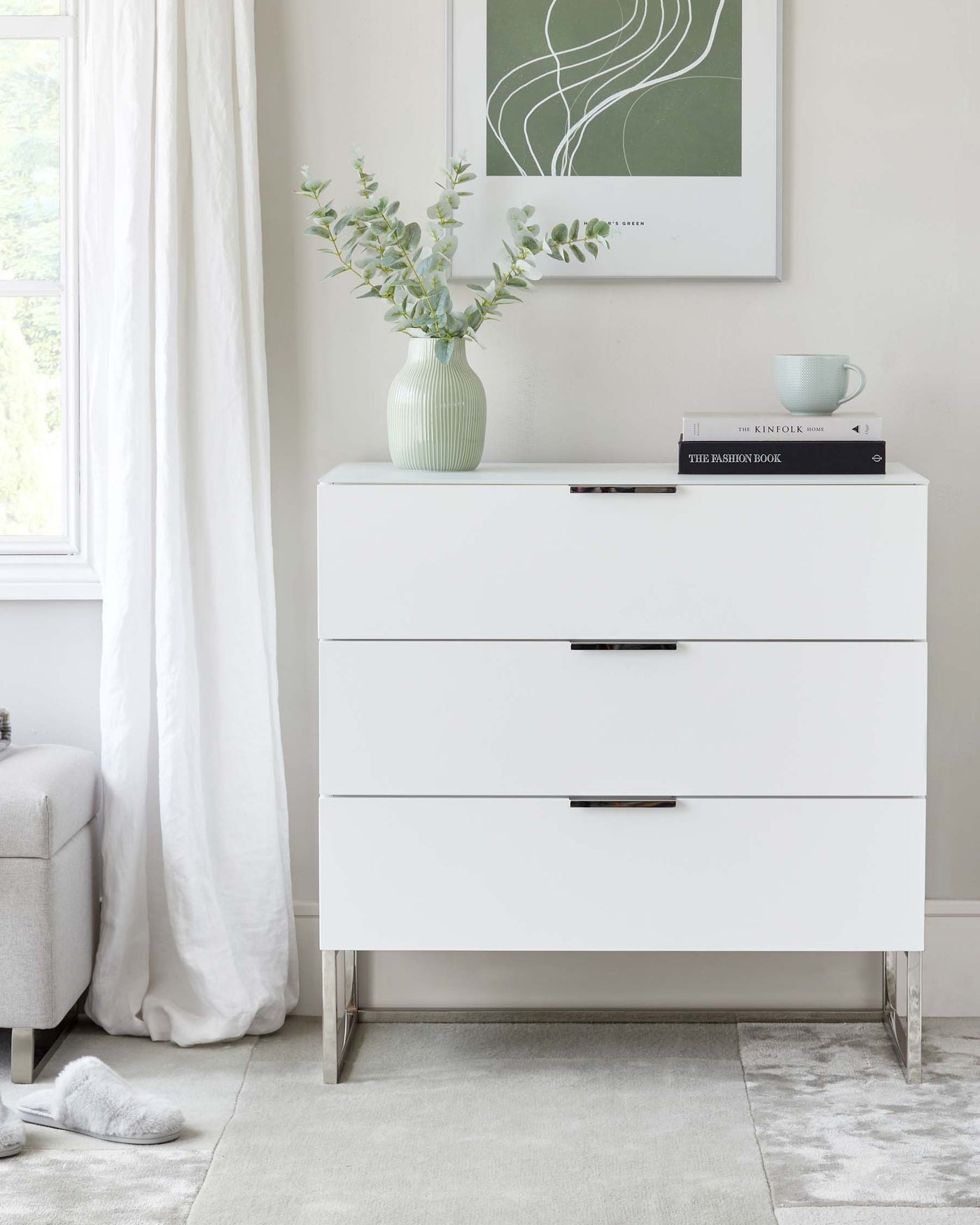 Modern white three-drawer dresser with sleek metal handles and minimalist metal legs, styled with a muted green vase with eucalyptus branches, two stacked books, and a light blue coffee cup on top, in a bright room with a grey upholstered bench, white curtains, and plush carpet flooring.