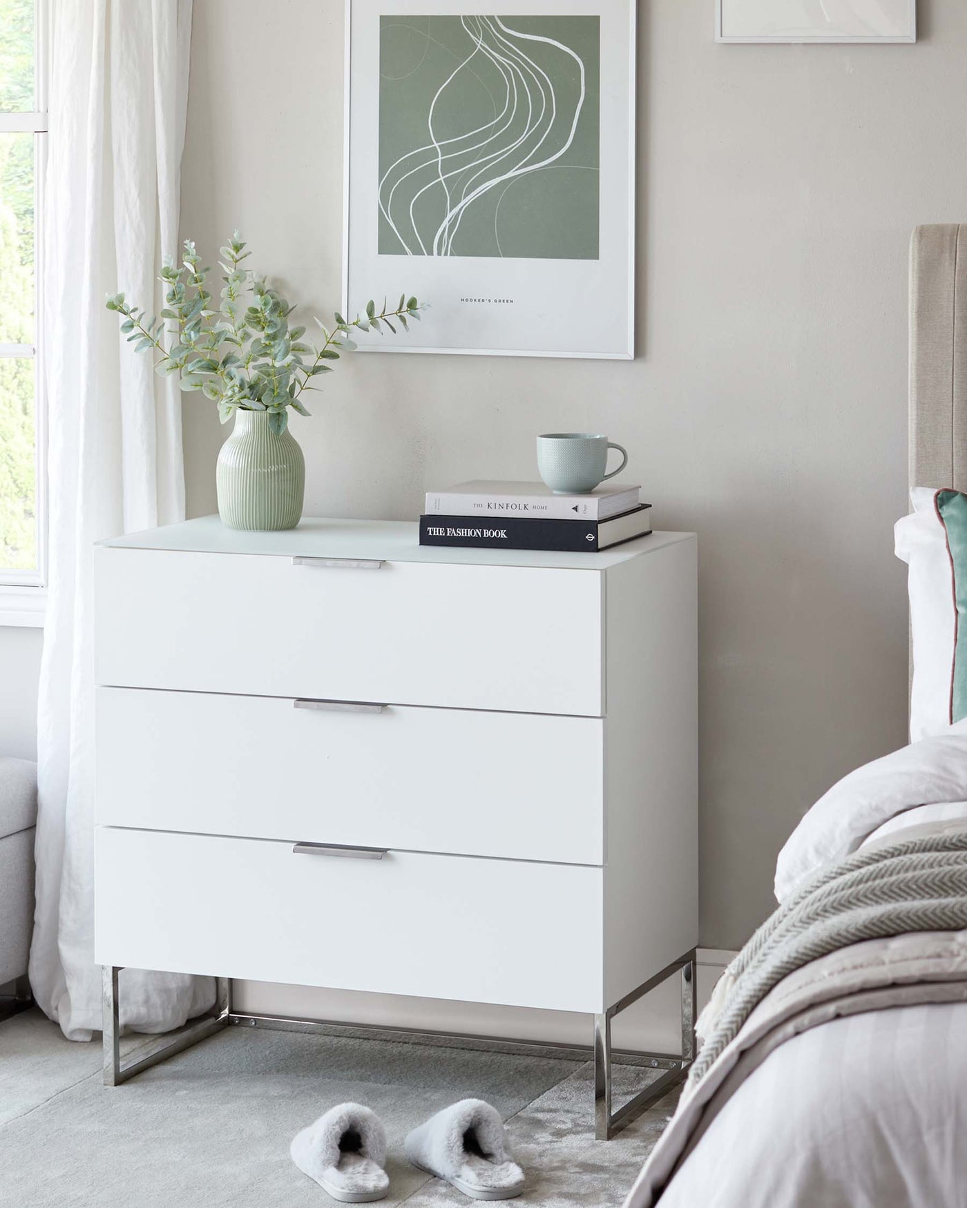 A modern white three-drawer chest with sleek handles and metal leg accents, situated in a softly lit bedroom setting.
