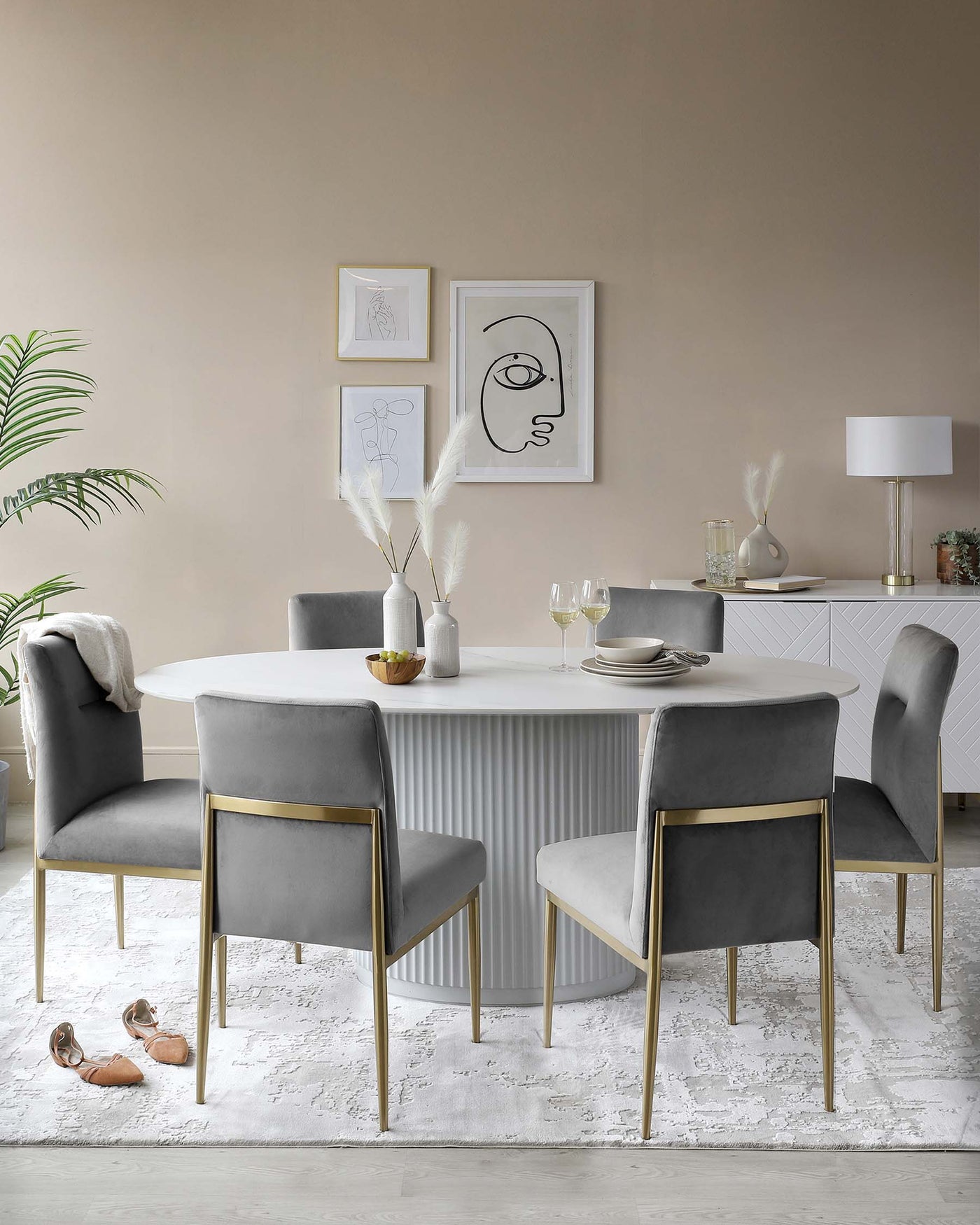 Elegant dining room featuring a round, white table with a pleated design on its pedestal base, accompanied by four plush velvet-upholstered chairs in a matching cool grey tone. Each chair sports a minimalist gold-finished metal frame, providing a sleek contrast against the soft upholstery. The furniture ensemble is set atop a subtle white textured area rug, adding a layer of warmth to the modern and sophisticated dining setting.
