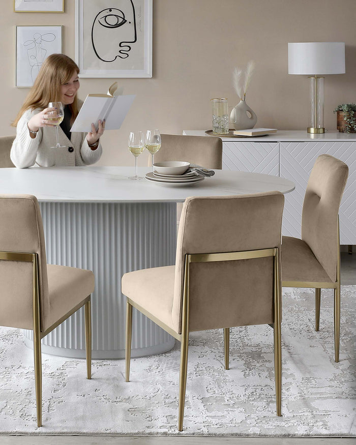 Elegant dining room setup featuring a modern, round white table with a fluted base, complemented by four plush, taupe upholstered chairs with sleek brass legs. A stylish white sideboard with geometric detailing and a contemporary white lamp with a cylindrical shade add to the sophisticated ambiance. The room is completed with a soft-textured white area rug beneath the dining set.