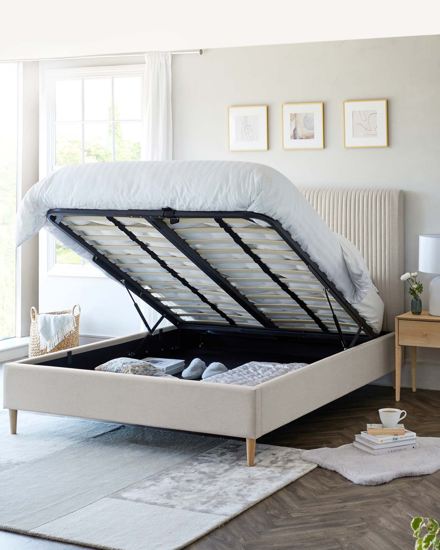 Contemporary light beige upholstered storage bed with a raised mattress platform revealing a spacious under-bed storage area, complemented by a light wood nightstand and a plush light grey area rug.