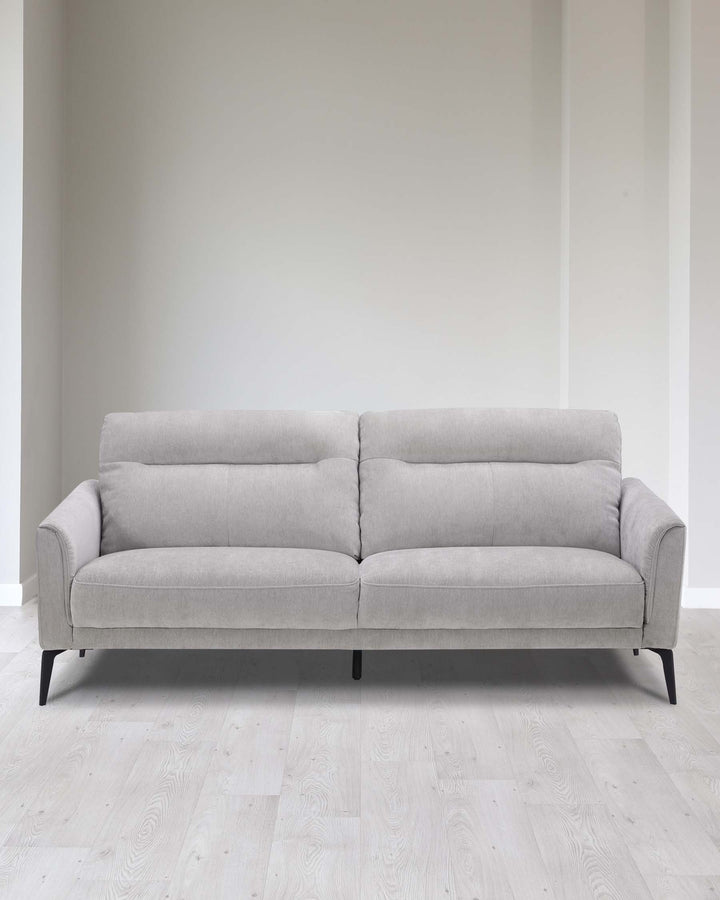 A modern light grey fabric sofa with a clean minimalist design, featuring a single row of tufting on the seat cushions, slightly flared armrests, and four black metal legs. The couch is set against a neutral backdrop with light wooden flooring.