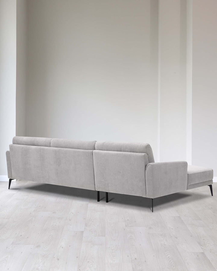 Elegant modern light grey fabric sectional sofa with clean lines, featuring a left-hand chaise lounge, straight arms, and black cylindrical legs, set in a minimalist room with light wooden flooring and white walls.