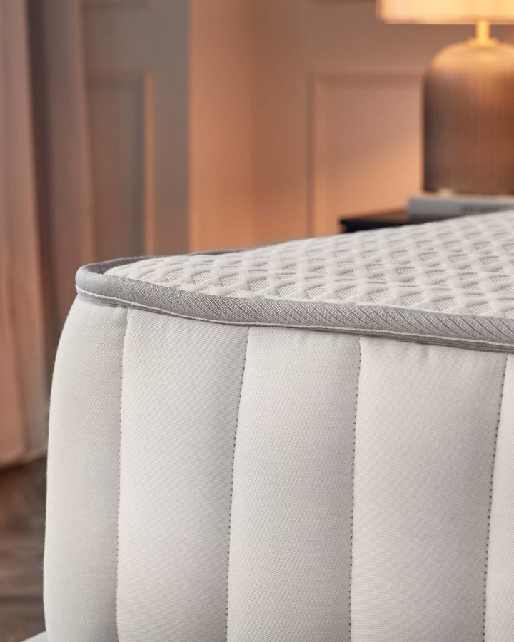 Elegant upholstered bench with textured light grey fabric, featuring piped edges and quilted detailing on the seating surface. In the background, there's a warm-toned wooden door and a metal lamp with a cylindrical shade, suggesting a cosy, contemporary interior setting.
