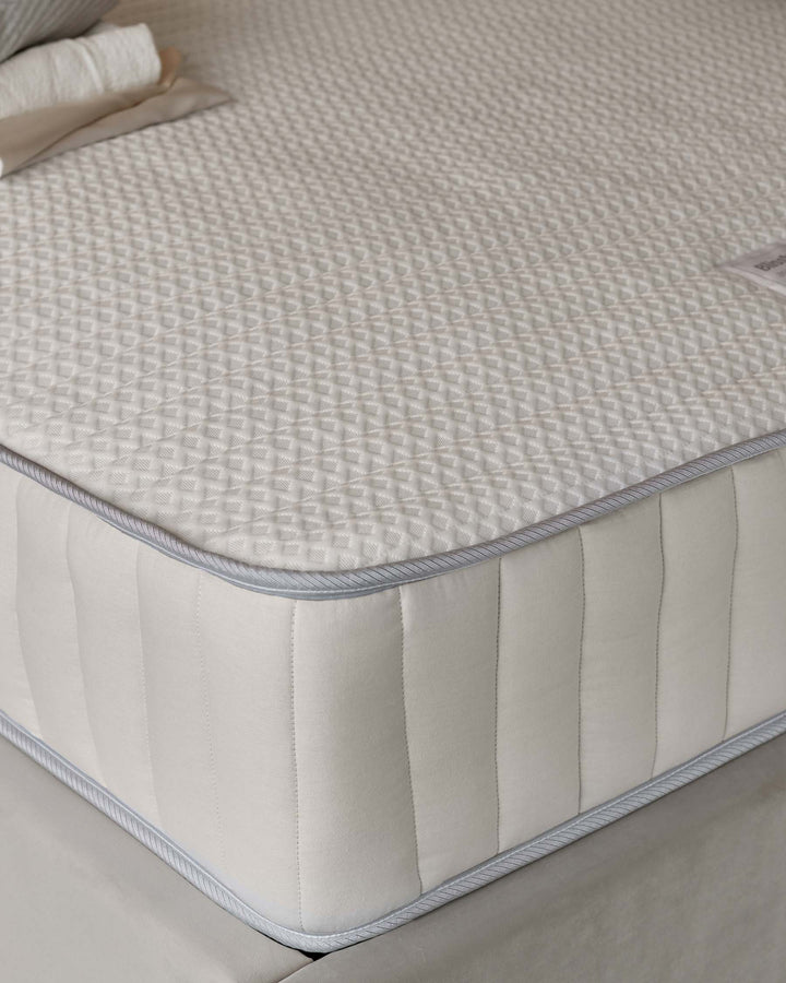 Close-up view of a premium-quality mattress featuring a quilted top with a diamond stitching pattern. The sides have vertically-stitched padding, and the mattress is edged with piping for a refined finish. A small section of a white pillow with a beige edge is also visible at the top corner, hinting at a coordinated bedding set.