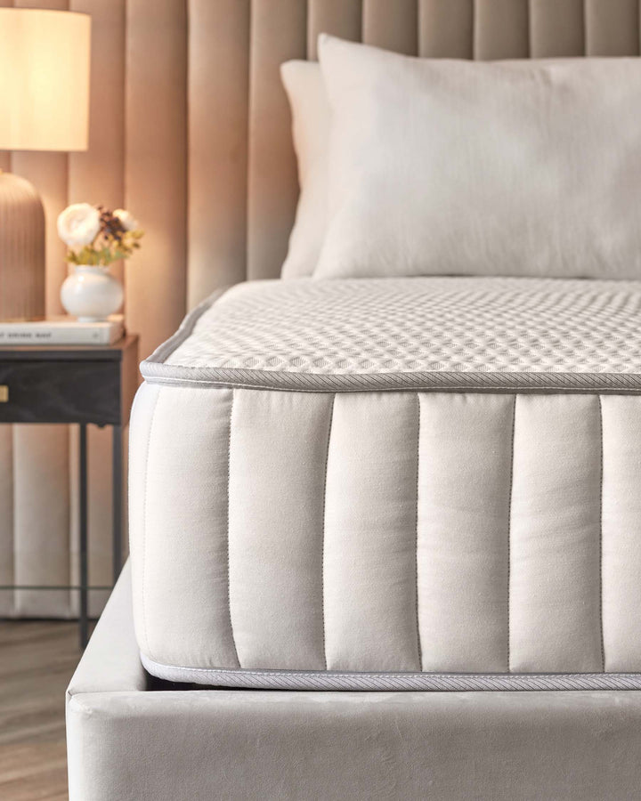 Close up of a mattress on a bed with a headboard and a bedside table with a lamp and flowers.