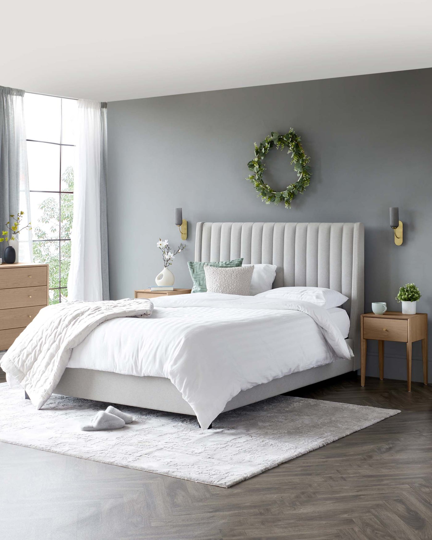 A modern bedroom featuring a large upholstered bed with a tall, tufted headboard. Beside the bed is a pair of mid-century-inspired nightstands with tapered legs. Opposite the bed stands a minimalistic, multi-drawer wooden dresser with simple knobs. The room is completed with a soft, textured area rug beneath the bed.