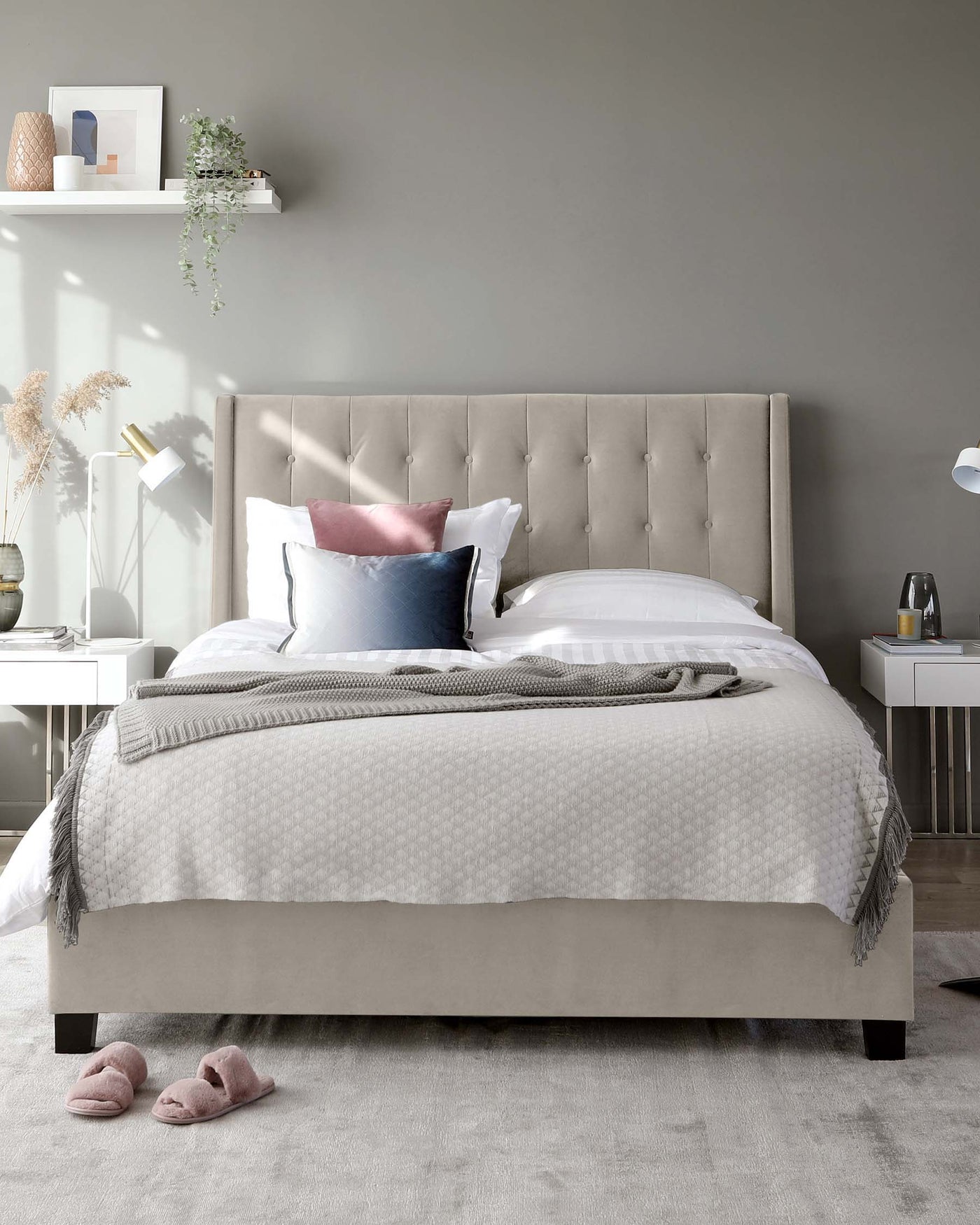 Elegant contemporary bed with a tufted headboard upholstered in a neutral grey fabric, complemented by a cosy bedding set with a mix of white and accent pillows, and a textured throw blanket. The bed is styled with symmetry in mind, flanked by matching white bedside tables and minimalist decor.
