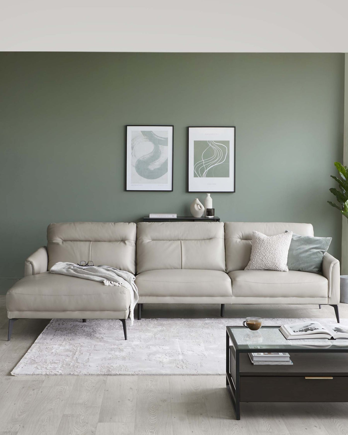 Modern light grey sectional sofa with chaise on the left side, featuring clean lines and plush cushions, positioned on an off-white distressed area rug. A rectangular black coffee table with a glass top and lower shelf sits in front of the sofa, accessorized with books and a cup.