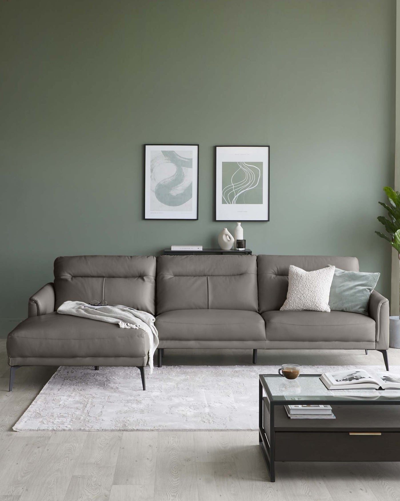 Modern L-shaped sectional sofa in a muted grey tone with clean lines and minimalistic metal legs, complemented by a sleek, rectangular black coffee table with a clear glass top and open shelf storage. A soft white area rug with a subtle pattern lies beneath the furniture ensemble.