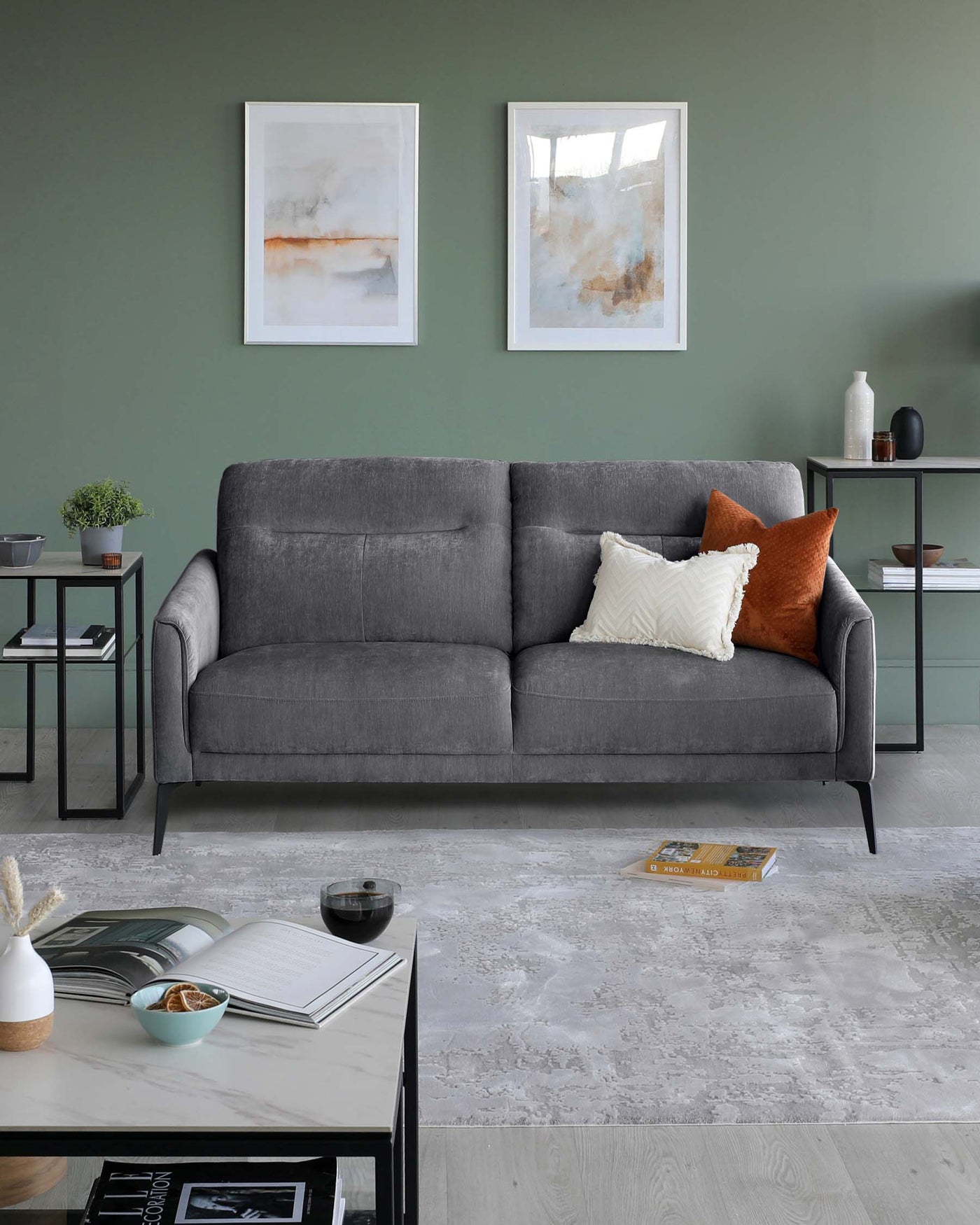 A modern living room featuring a charcoal grey fabric sofa with clean lines and slender black metal legs, complemented by a mix of geometric and textured throw pillows. In front of the sofa stands a sleek black metal side table with a square top, accessorized by a small potted plant, beside a matching minimalist rectangular coffee table displaying books and decorative items on both the top and lower shelf.