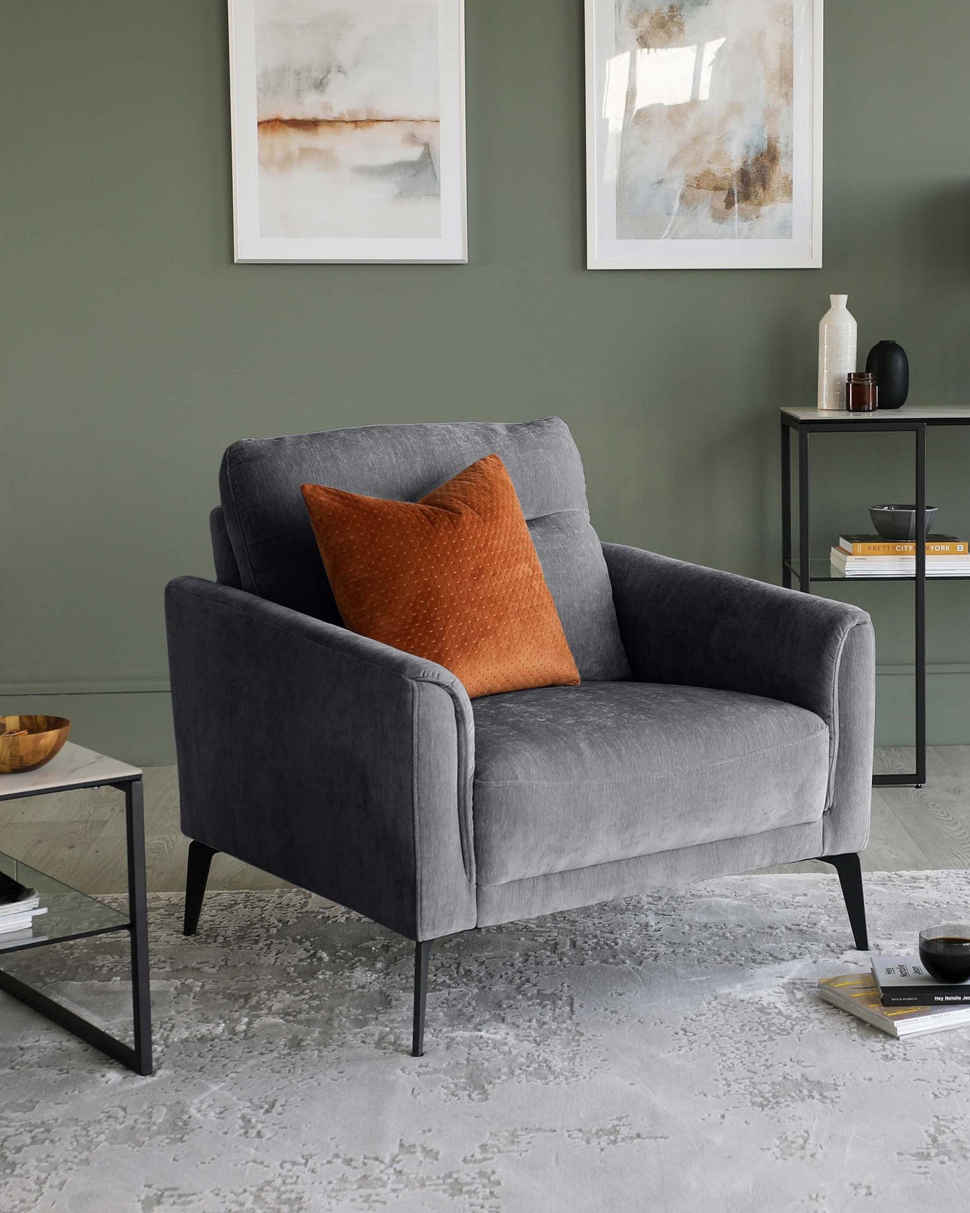Elegant contemporary-style armchair with a smooth, grey upholstery and a single burnt orange textured throw pillow. The armchair features sleek lines, slightly flared armrests, and slender black metal legs. To the right, there's a minimalist black metal frame side table with a rectangular glass top, holding decorative items and books. The furniture is set against a muted green wall and atop a distressed grey area rug, complemented by abstract wall art.