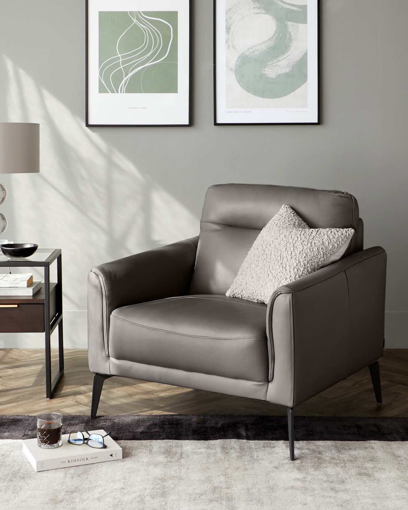 Modern grey leather armchair with a textured throw pillow, complemented by a sleek black side table with a round top, situated on a grey and black area rug against a wall adorned with framed abstract art pieces.