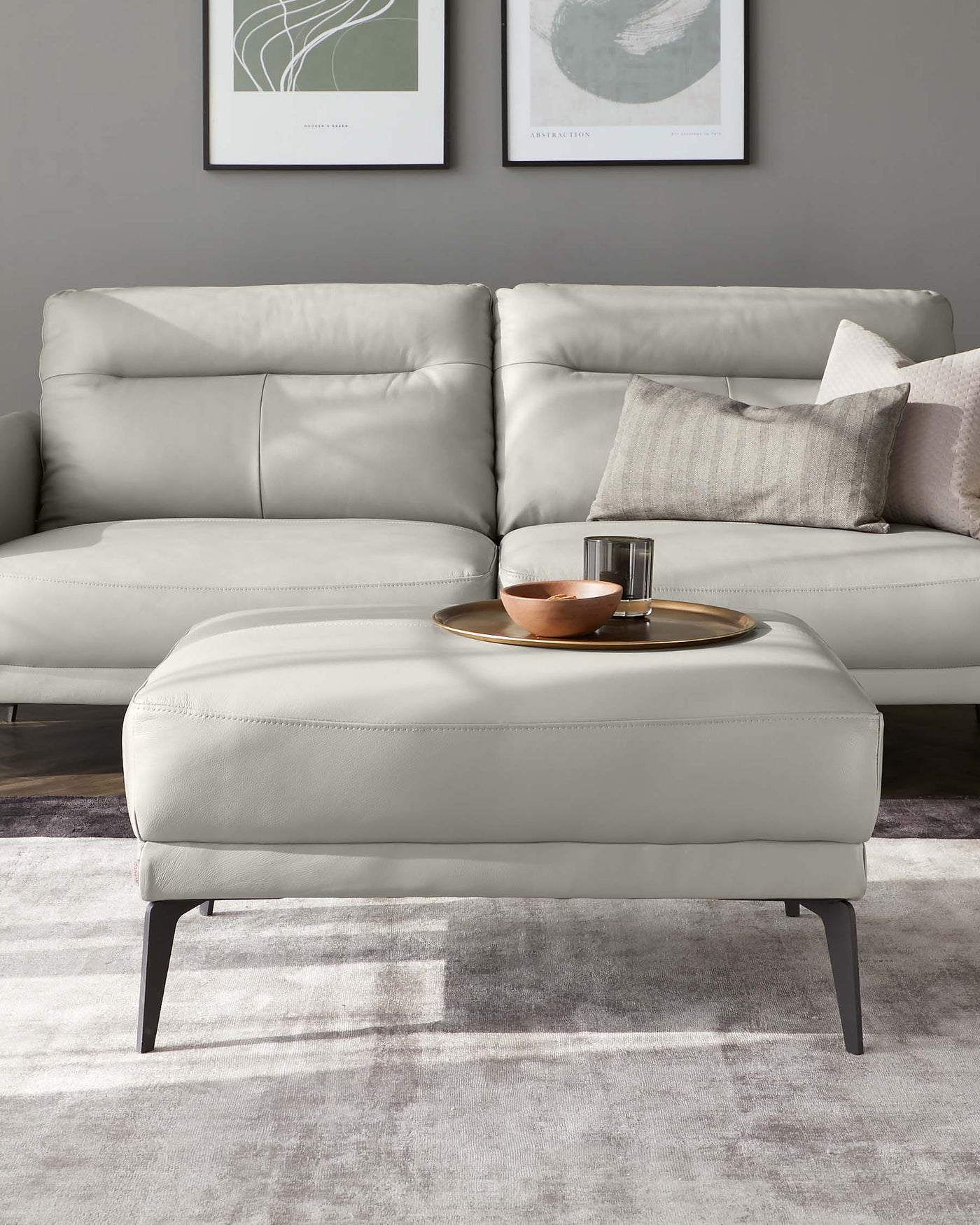 Modern light grey leather sectional sofa with plush cushions and a matching rectangular ottoman featuring a minimalist black metal leg design. The ottoman is accessorized with a round tray holding a decorative copper bowl and a clear glass. The sofa is set against a grey wall with abstract framed art, placed on a gradient grey area rug.