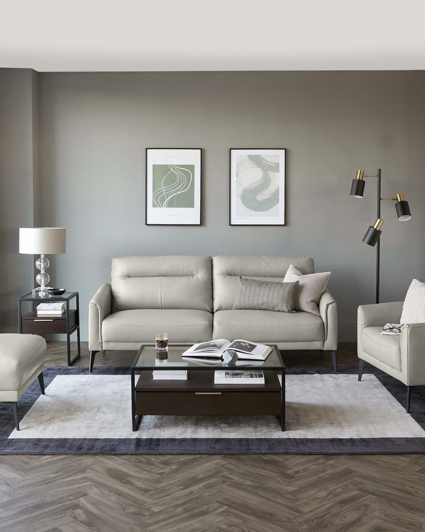 A modern living room setup featuring a light grey upholstered sofa with matching armchair and ottoman. A sleek, rectangular wooden coffee table with a glass top sits on a textured white and grey area rug, accompanied by a dark wood side table with a drawer. A tall floor lamp with multiple adjustable arms is positioned in the corner. Two framed abstract artworks hang above the sofa, completing the contemporary aesthetic.