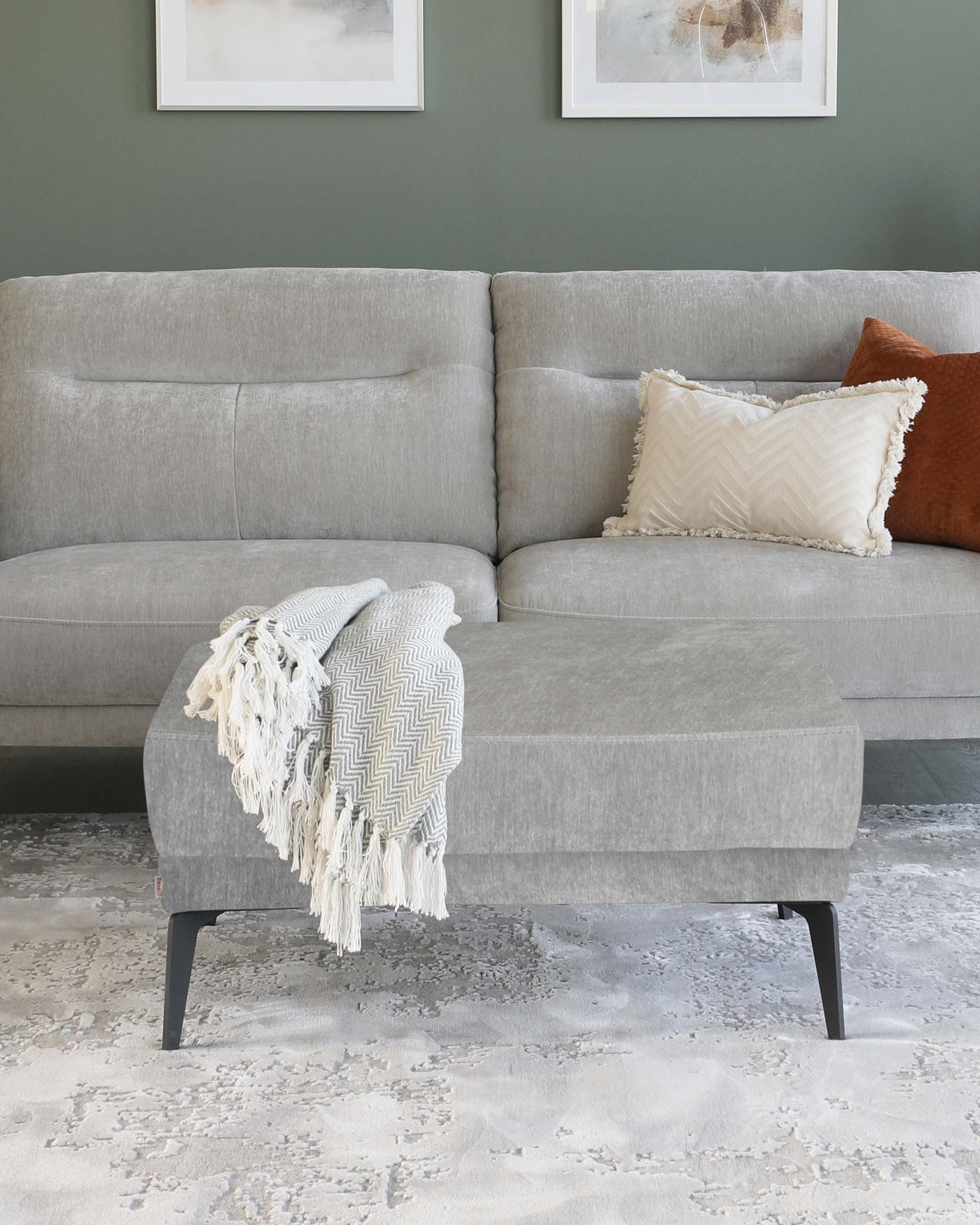 Contemporary grey sectional sofa with textured upholstery and plush cushions, complemented by a matching grey ottoman with an angular black metal base. Accents include a cream fringed throw and decorative pillows in earth tones.