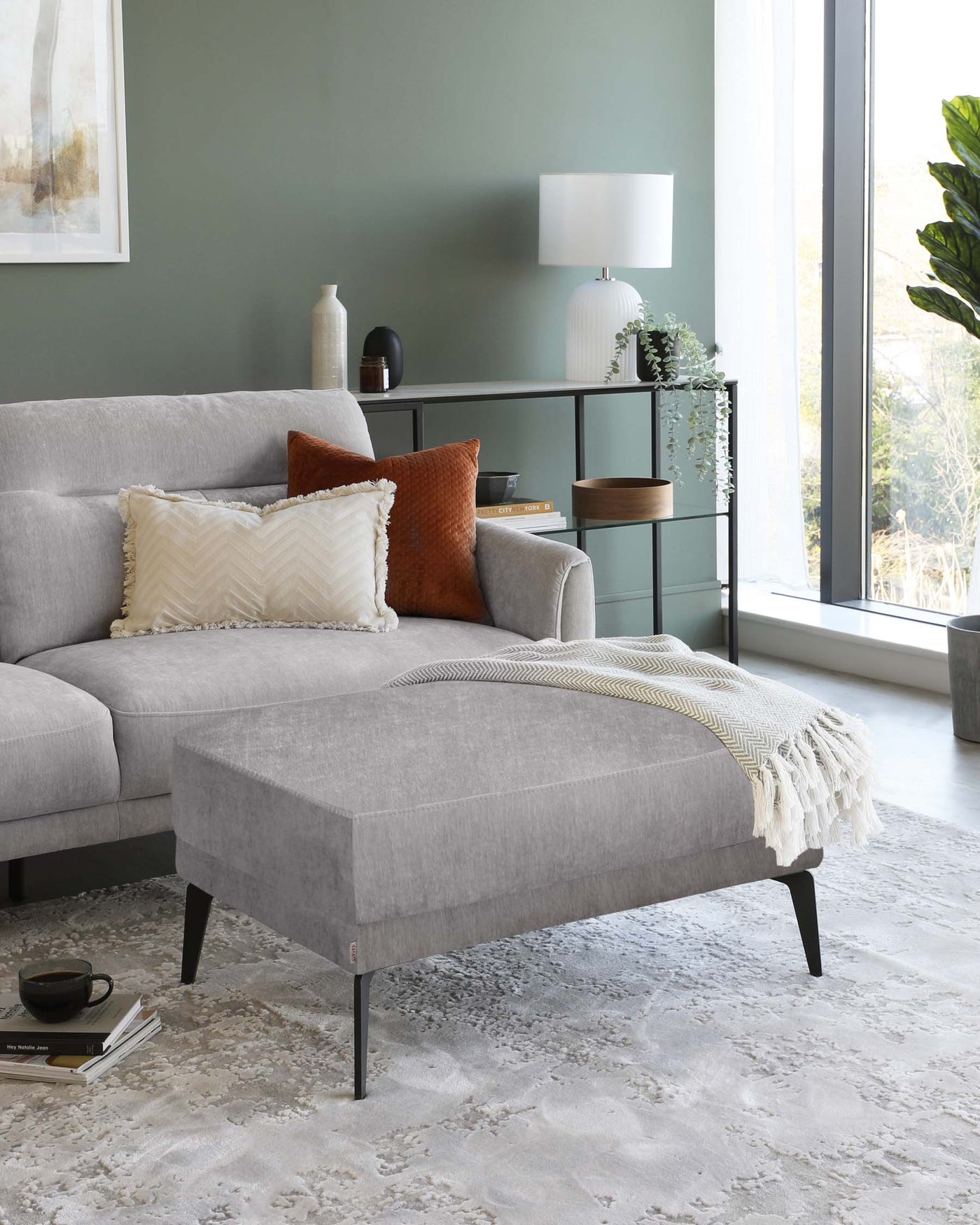Modern grey fabric sofa with plush cushions and a coordinating grey fabric ottoman featuring dark tapered legs. A sleek black metal and wood shelving unit with decorative items is visible in the background. The furniture arrangement is on a textured off-white area rug, contributing to a contemporary and comfortable living space aesthetic.