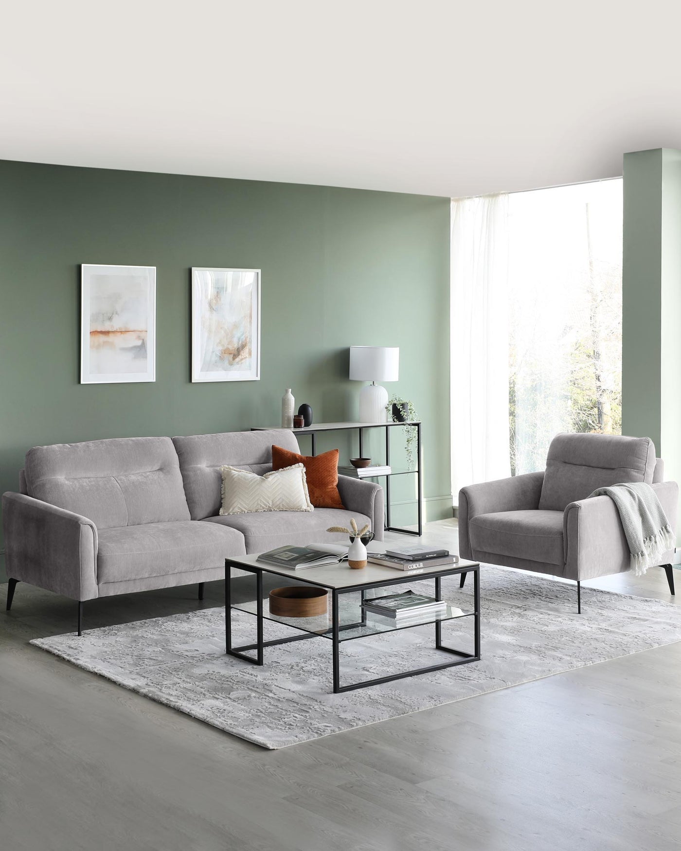 Modern living room furniture featuring a grey fabric three-seater sofa and a matching armchair with sleek metal legs, accompanied by a set of two black metal-framed coffee tables with glass tops, and a coordinating metal-framed side table with a glass top and a lower shelf. A soft grey area rug anchors the arrangement on a light wood floor.