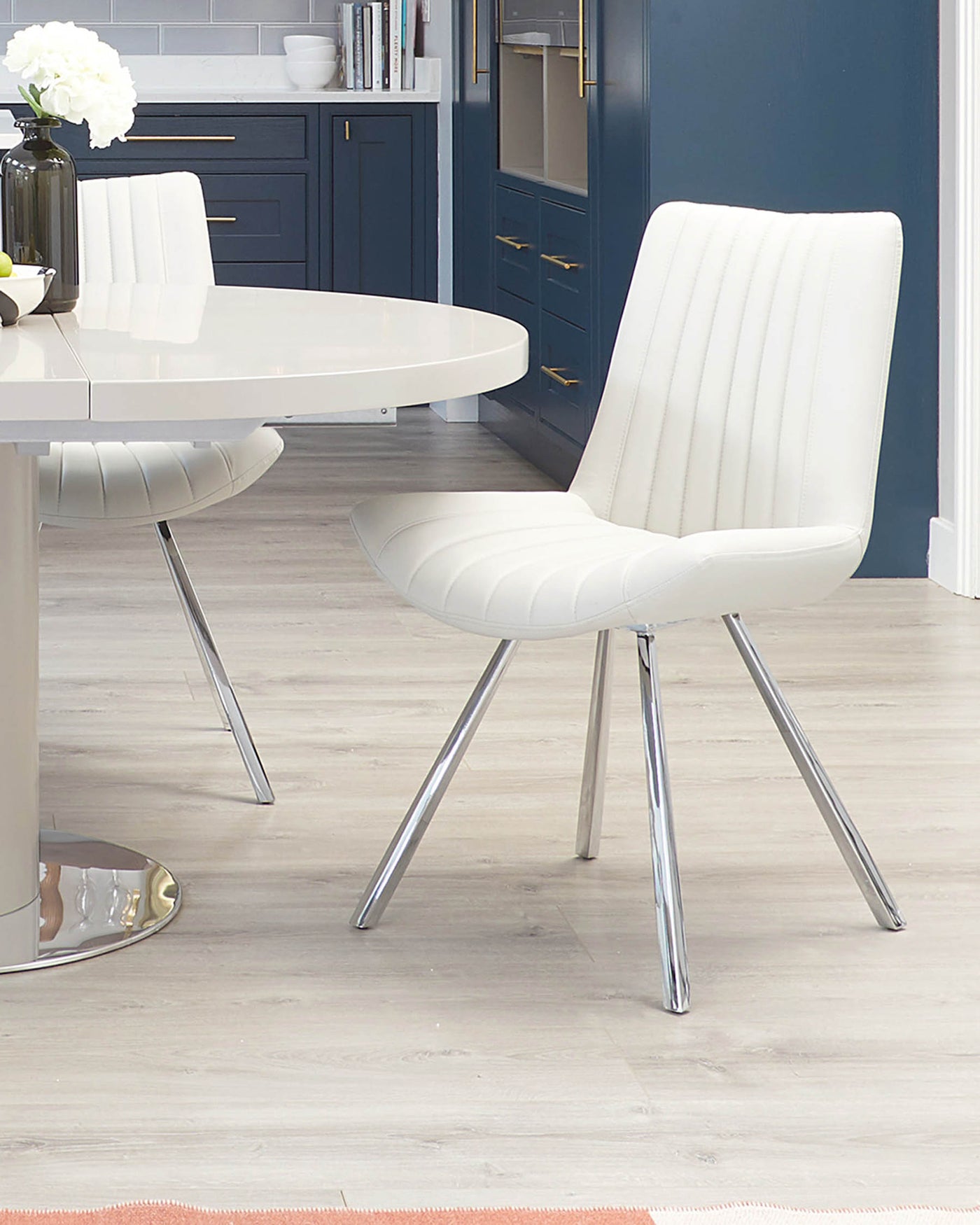 Modern white oval dining table with a sleek, reflective surface, complemented by two elegant white dining chairs featuring horizontal channel tufting and polished chrome legs. The chairs have a curved, shell-like seat design that provides both comfort and contemporary aesthetic.