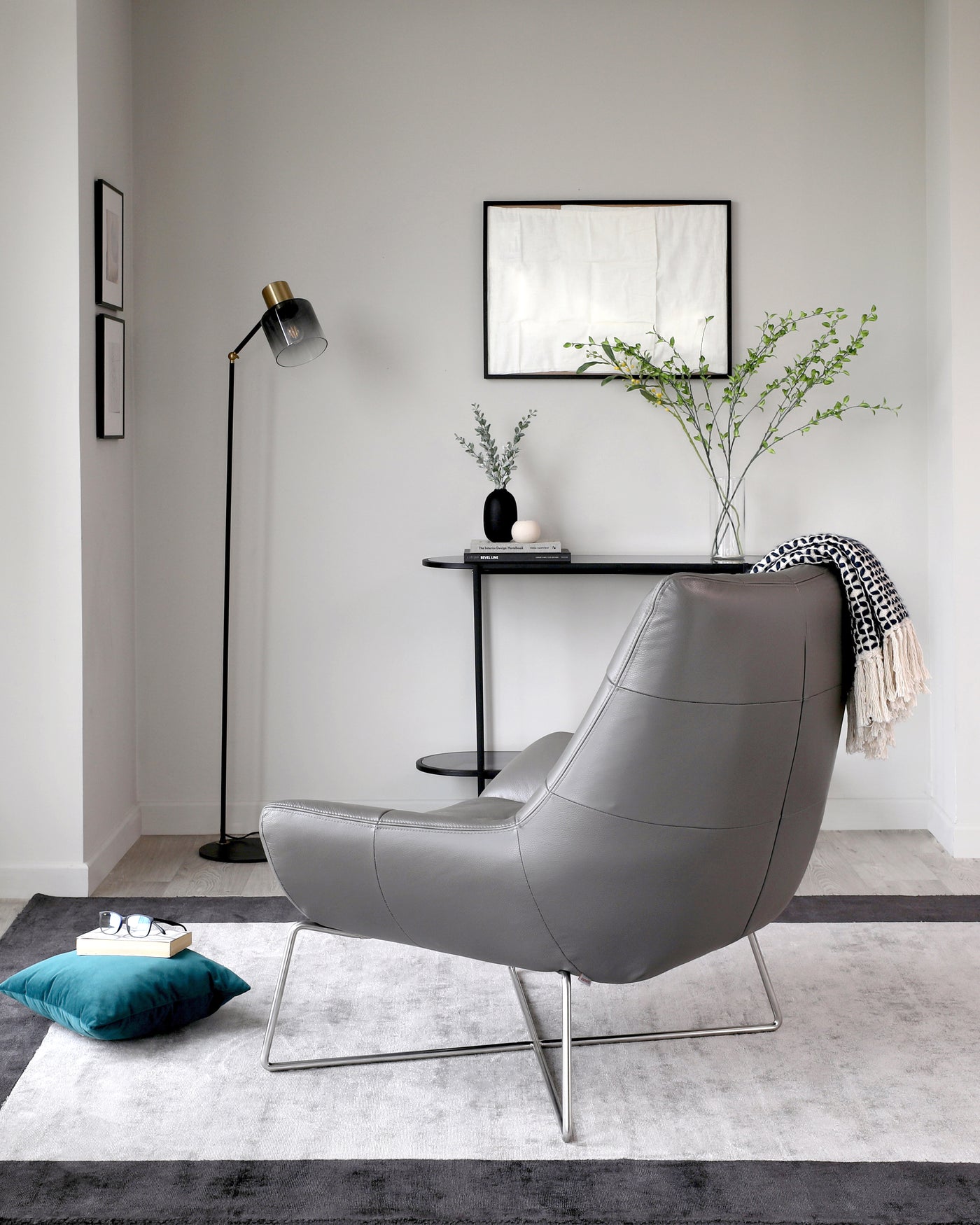A modern grey upholstered lounge chair with a unique curved design and sleek metal legs sits on a two-tone grey area rug. Behind it, a round black side table holds a vase and book, accented by a tall floor lamp with a glass shade to the left. A framed abstract artwork hangs on the wall above the table, adding a touch of sophistication to the minimalist decor.