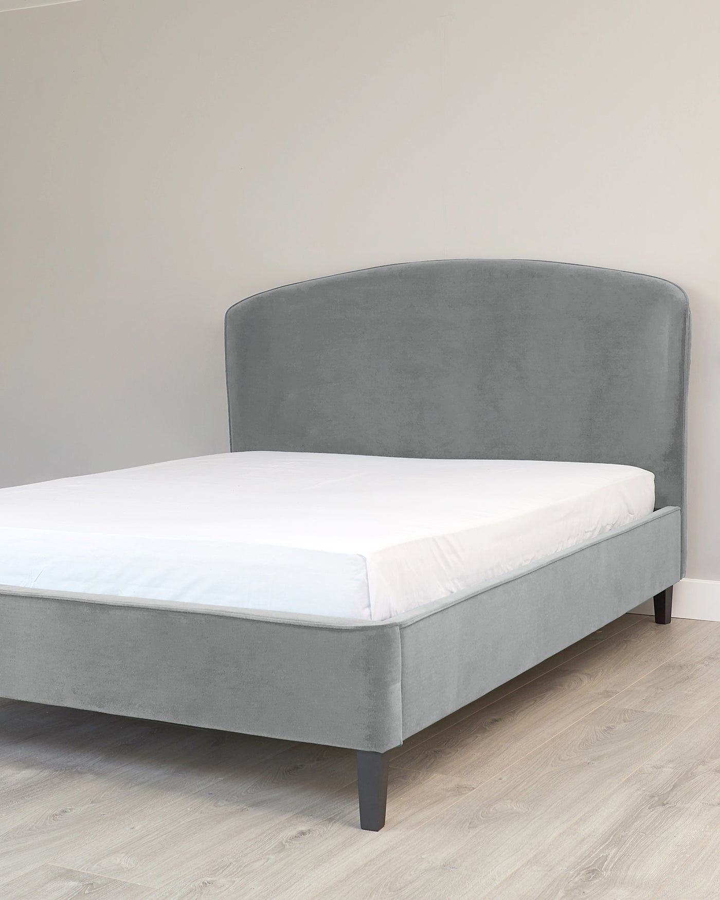 Grey upholstered platform bed with a curved headboard and simple dark wooden legs, showcased in a room with light wooden flooring and a neutral wall. A white mattress is placed on top of the bed frame.