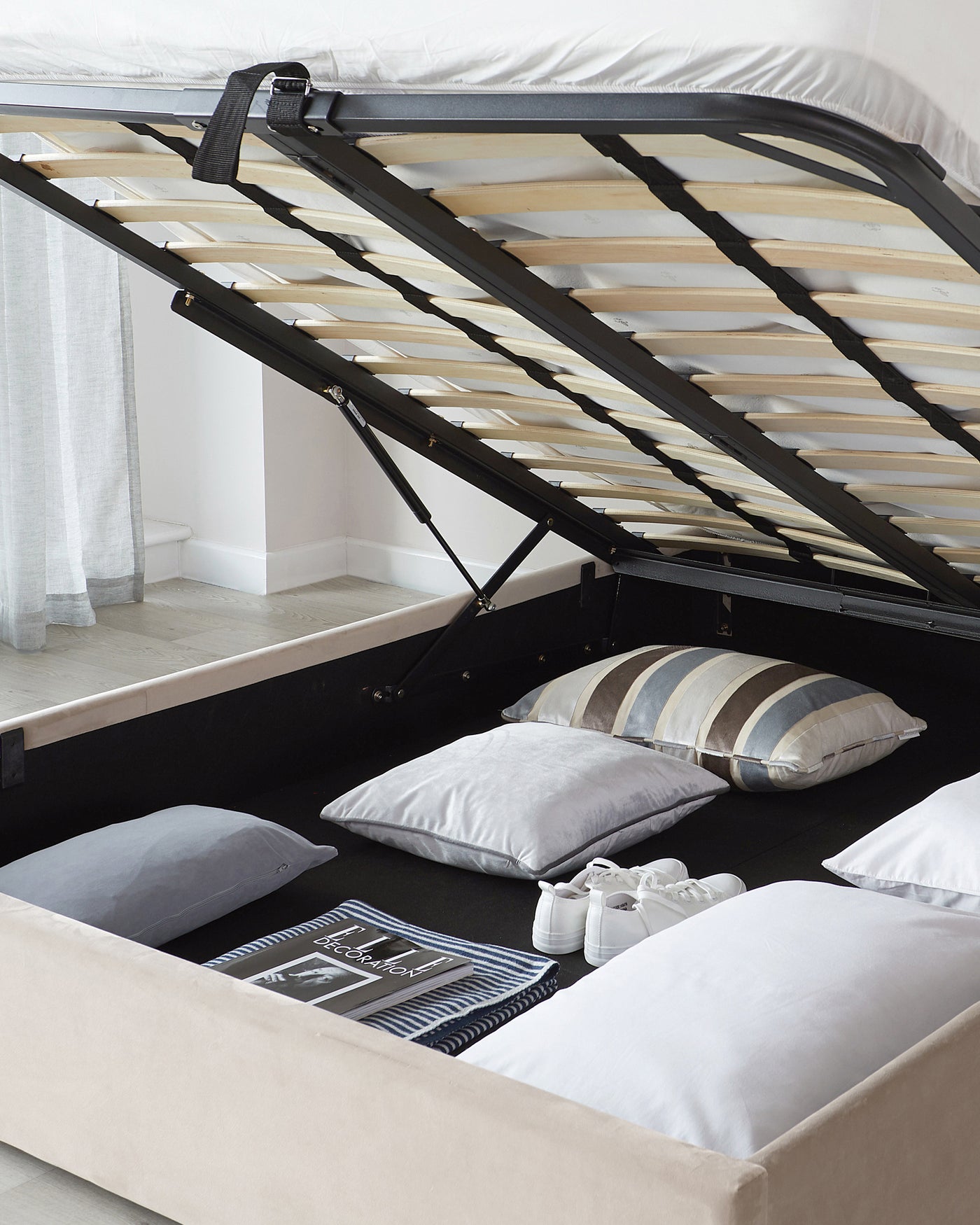 Contemporary upholstered storage bed with a lifting frame revealing a spacious storage compartment underneath. The bed features a beige fabric finish and a slatted wood mattress support which is elevated, with a black support mechanism visible. Inside the storage space are various pillows and a magazine, indicating the bed's utility for storing bedding and personal items.