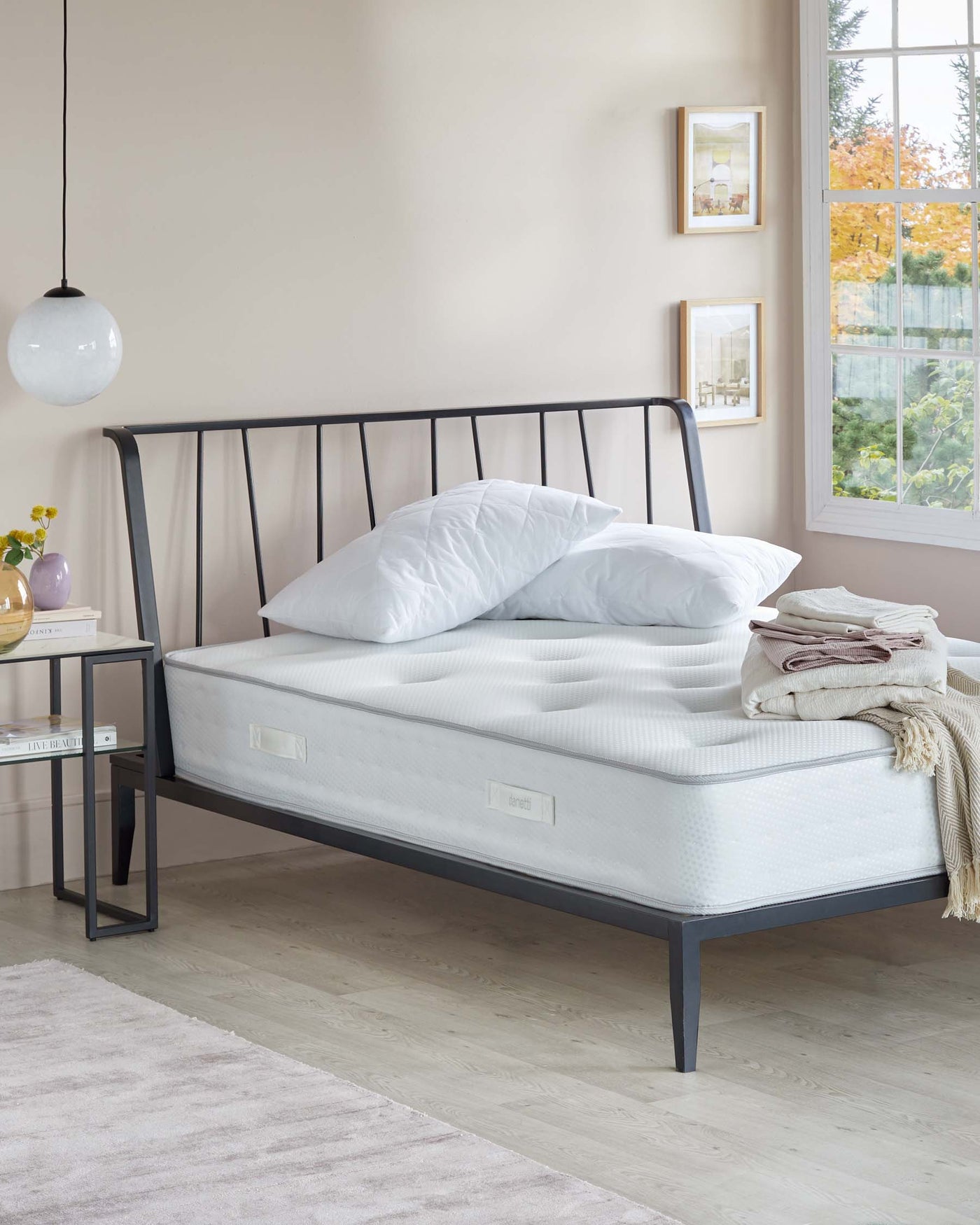 Elegant minimalist bedroom with a contemporary metal bed frame in black finish and a comfortable white mattress. Beside the bed is a modern black metal side table with a simple square design, displaying books and decorative items.