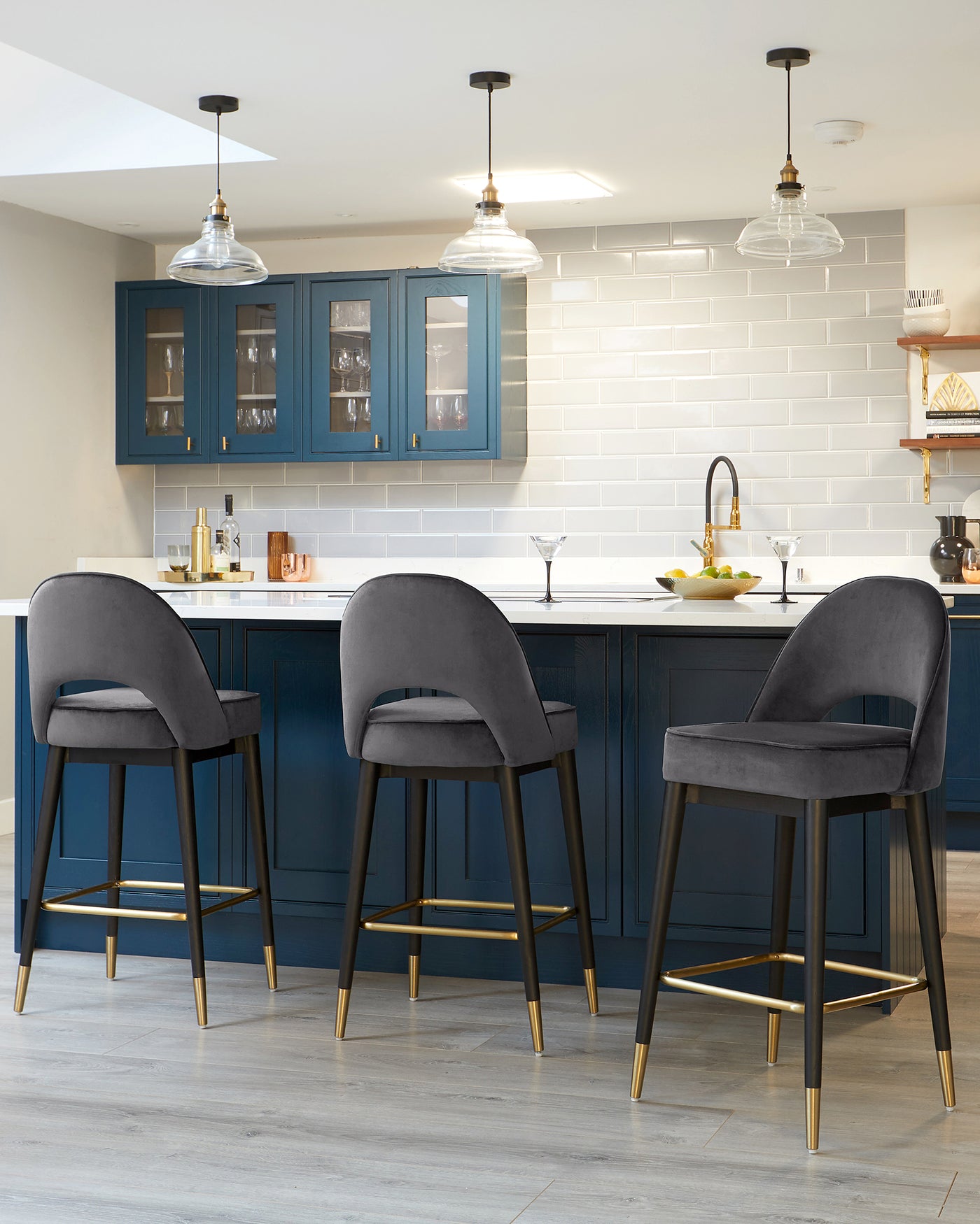 Three contemporary bar stools with plush grey upholstery and black frames accented with gold footrests, positioned at a kitchen island.