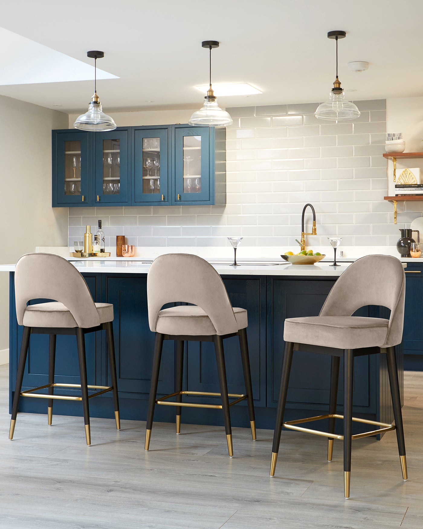 Three elegant, contemporary bar stools with plush, light grey upholstery and black wooden frames featuring brass-finished accents at the base of the legs and footrests.