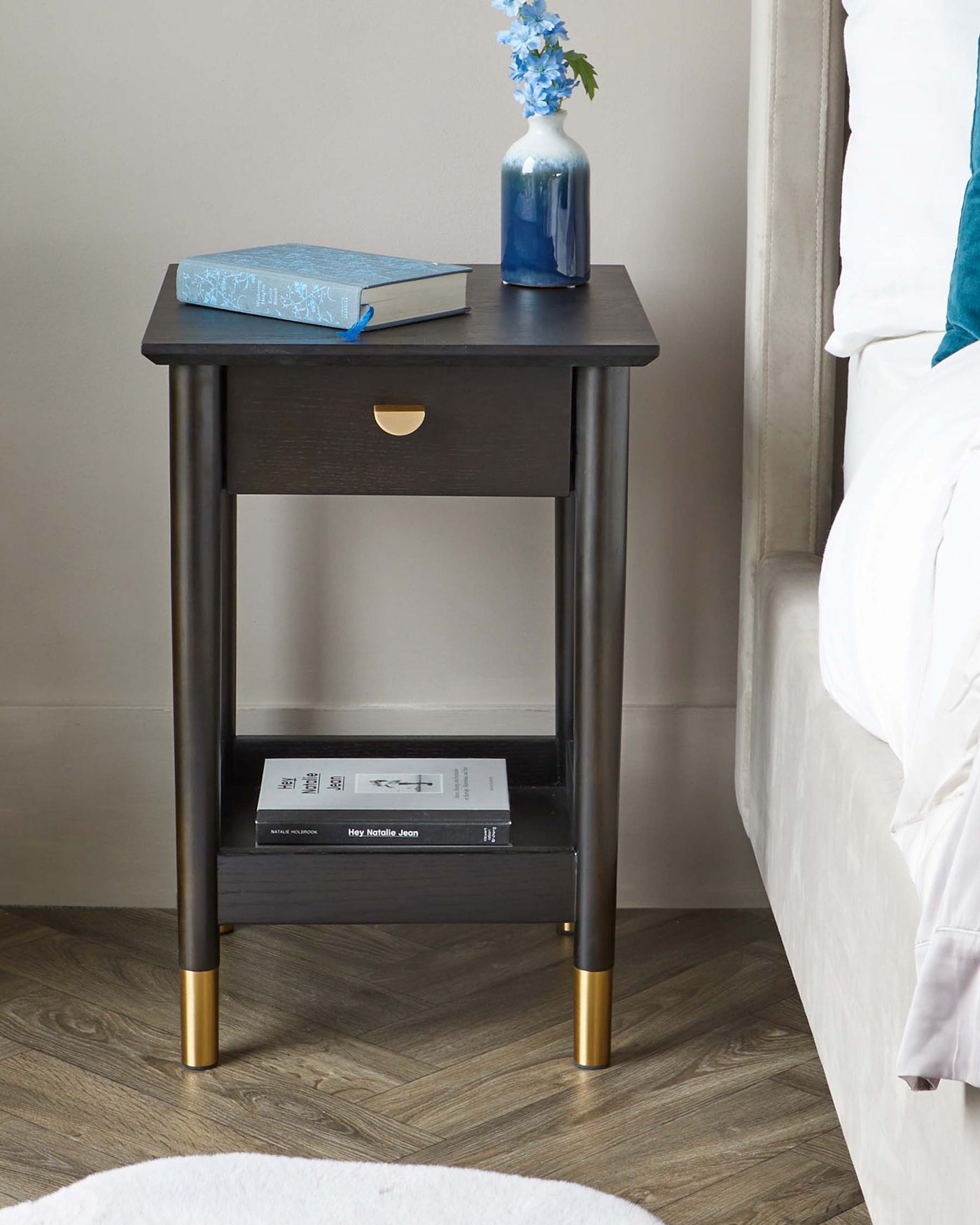 A contemporary espresso-finished nightstand featuring a smooth rectangular top, a pull-out drawer with a brass crescent-shaped pull, and tapered legs with brass caps. The lower shelf offers additional storage space, and the piece is styled with a book and a vase of flowers on top.