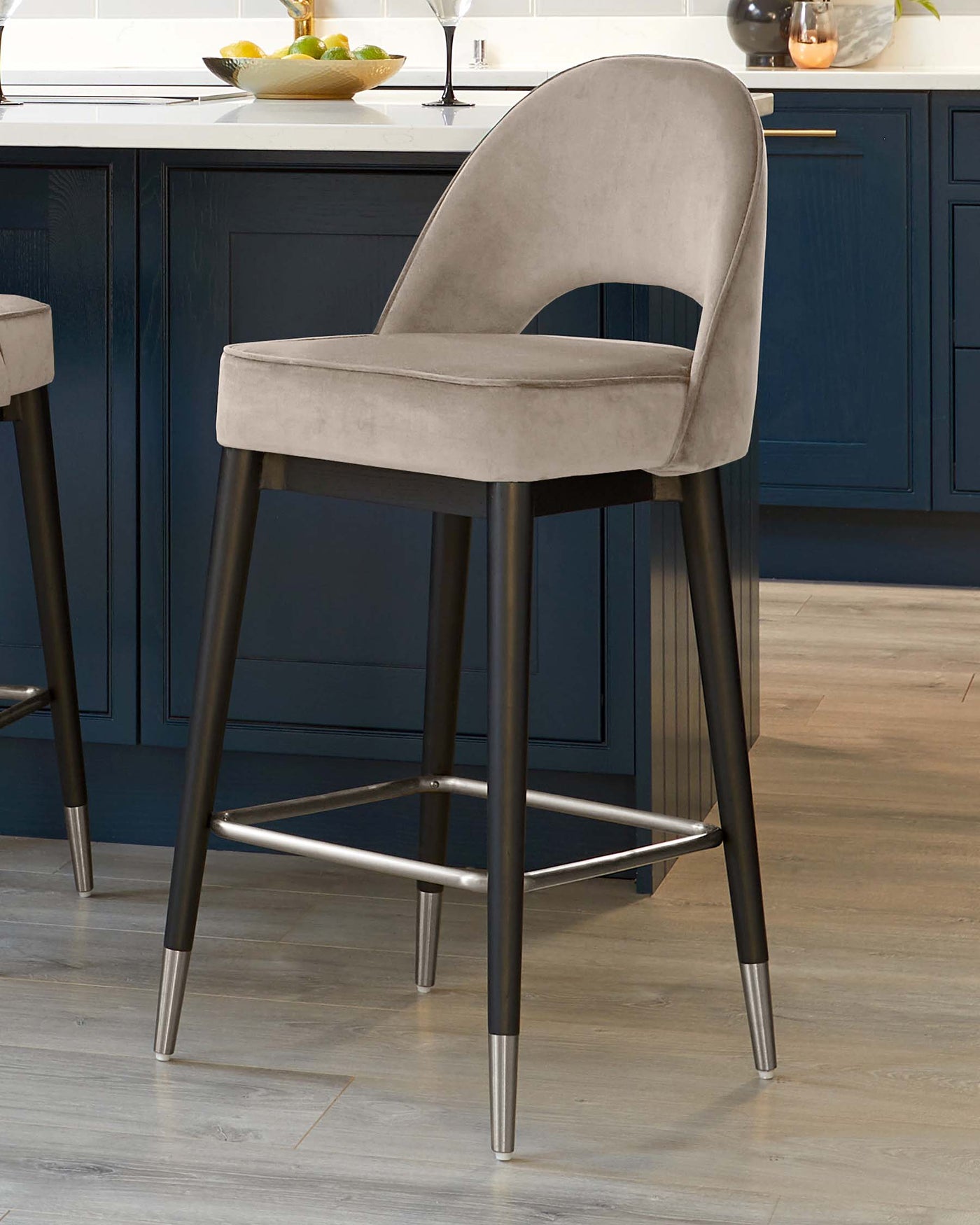 Elegant modern barstool with velvety taupe upholstery and a curved seat back, featuring black tapered legs with metallic footrest and tips.