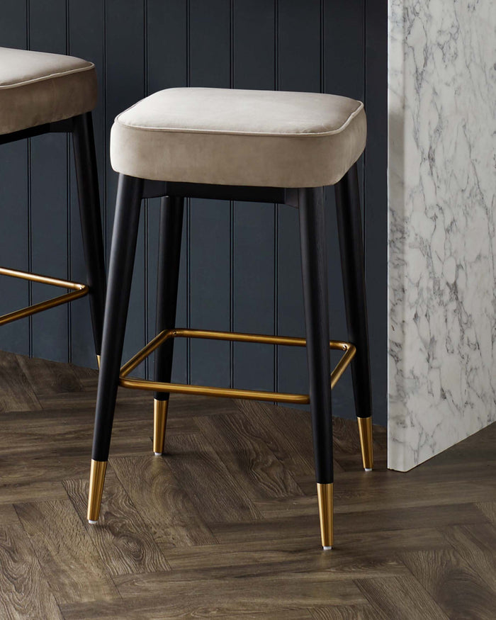 Elegant modern bar stools with plush, taupe upholstered seats and sleek black frames accented with gold-finished footrests and leg tips, displayed against a dark wood floor and panelled wall.