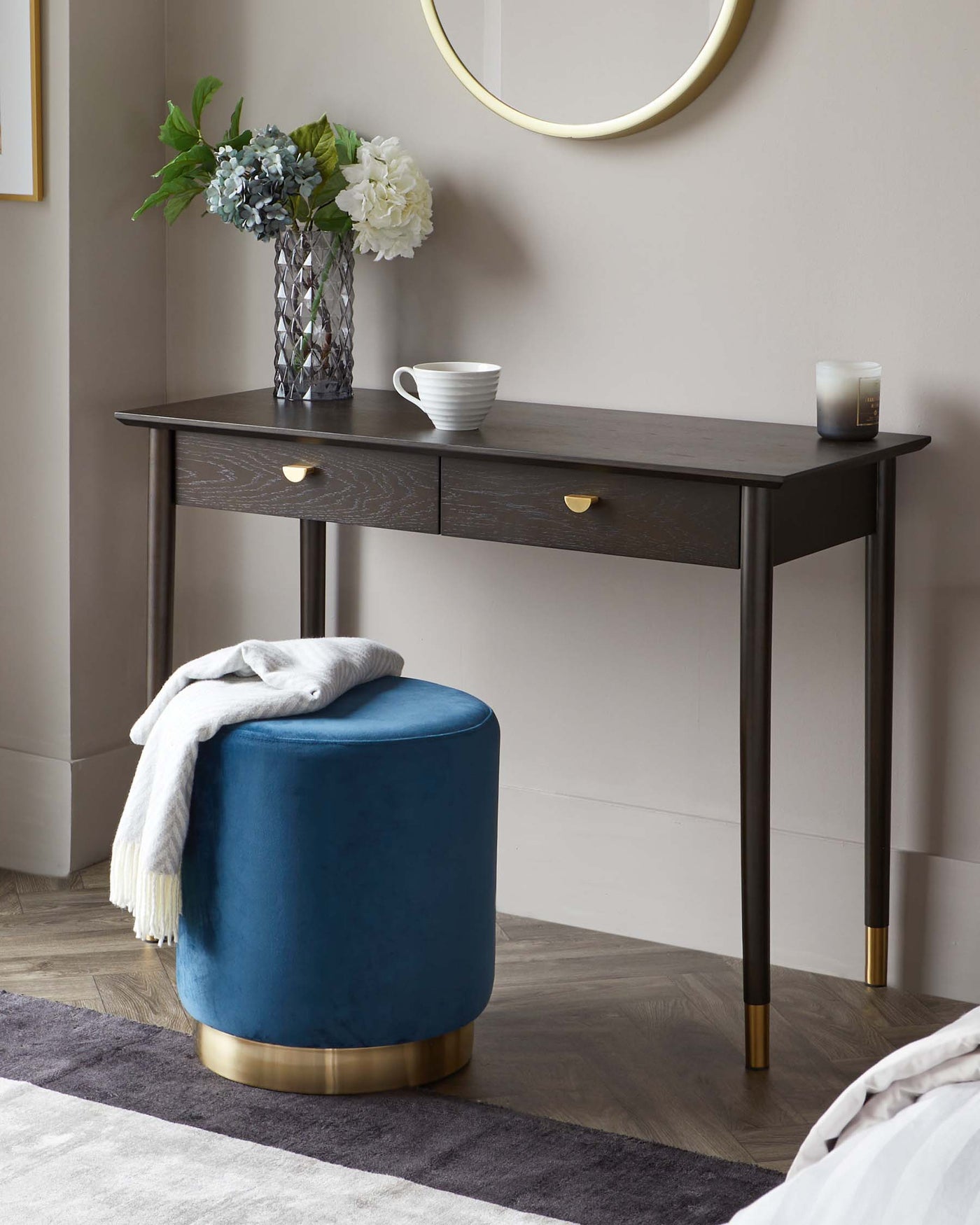 A dark wood console table with two drawers, featuring tapered legs with gold-toned tips, accented by brass half-moon drawer pulls. Paired with the table is a round blue velvet ottoman with a gold-toned metallic base, adding a touch of luxury to the setting.