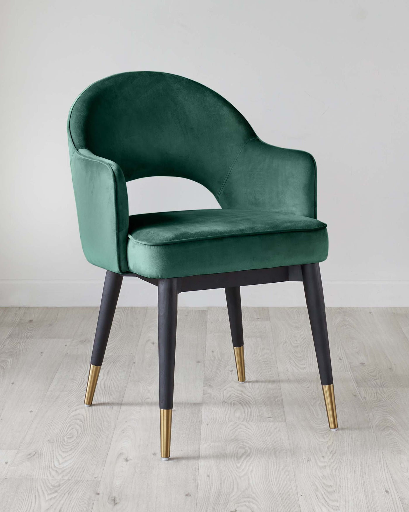 Elegant modern armchair with dark green velvet upholstery and matte black legs tipped with brass accents, set against a light grey wooden floor and white wall background.