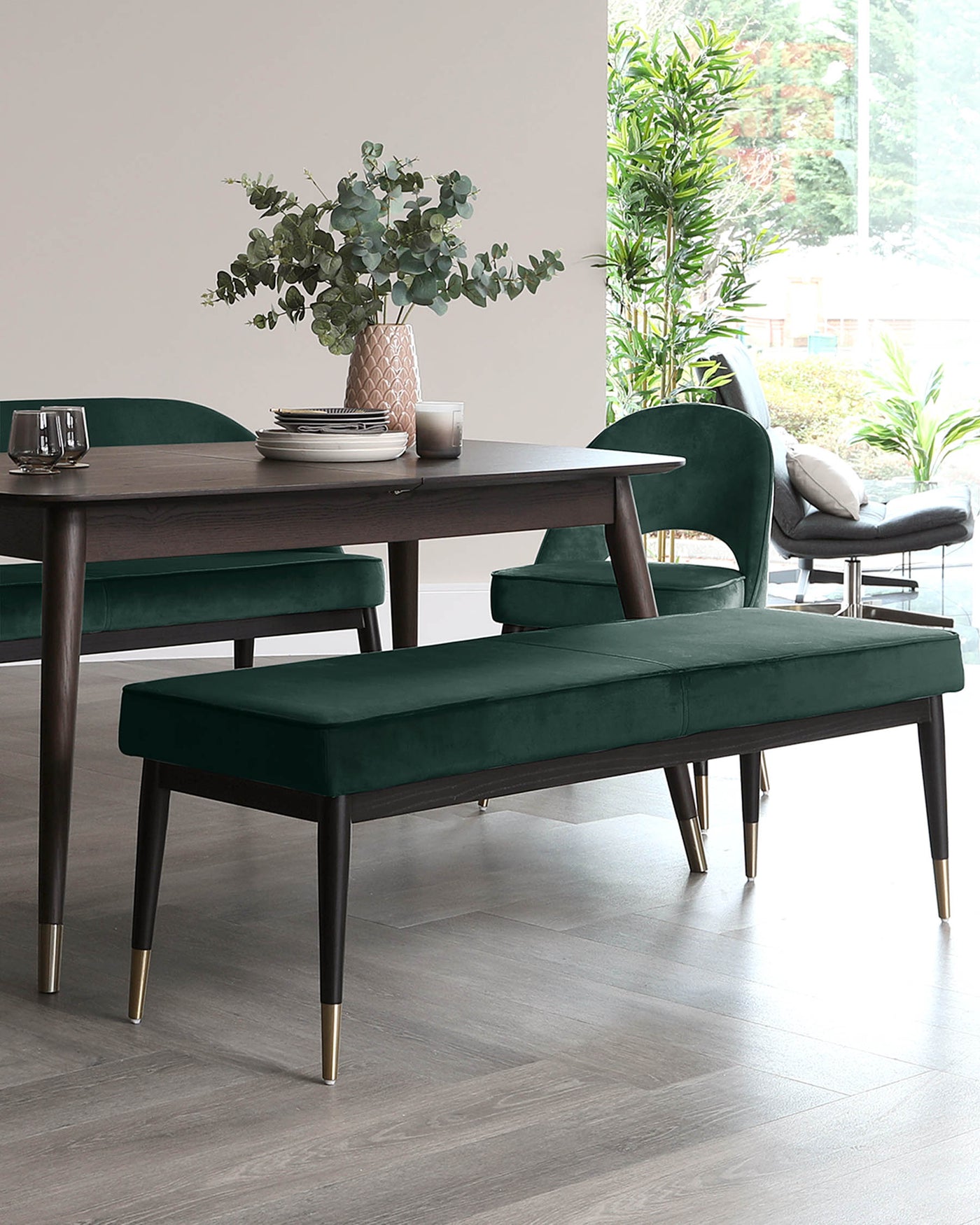 A dining set featuring a dark wood table with an elegant, minimalistic design and tapered legs. Two plush, emerald green velvet upholstered benches with dark wooden legs and brass detailing serve as seating, with one bench tucked neatly under the table.