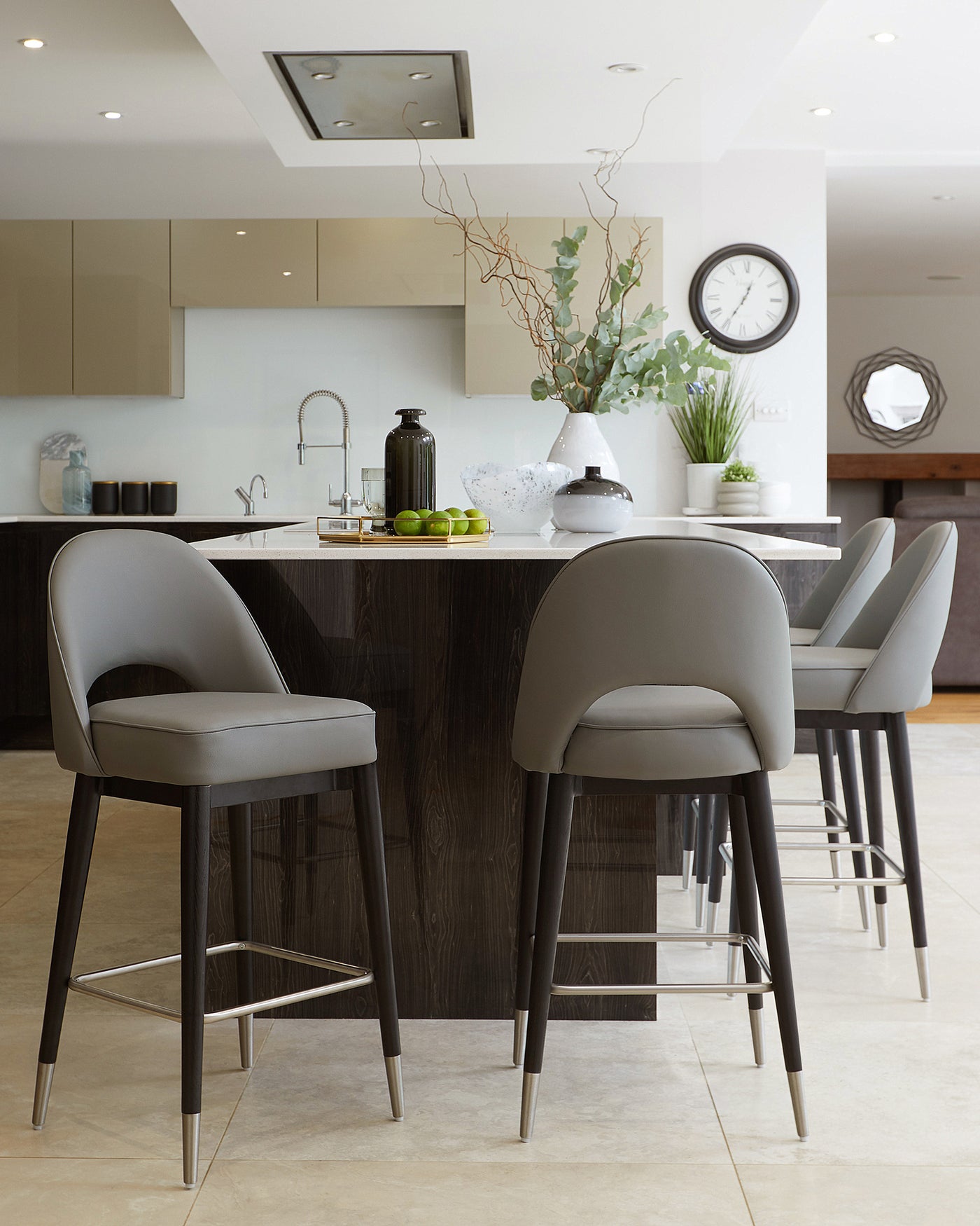 Modern kitchen interior with three elegant grey upholstered bar stools featuring slender black legs with metallic footrests, positioned at a dark wood kitchen island with a white countertop.