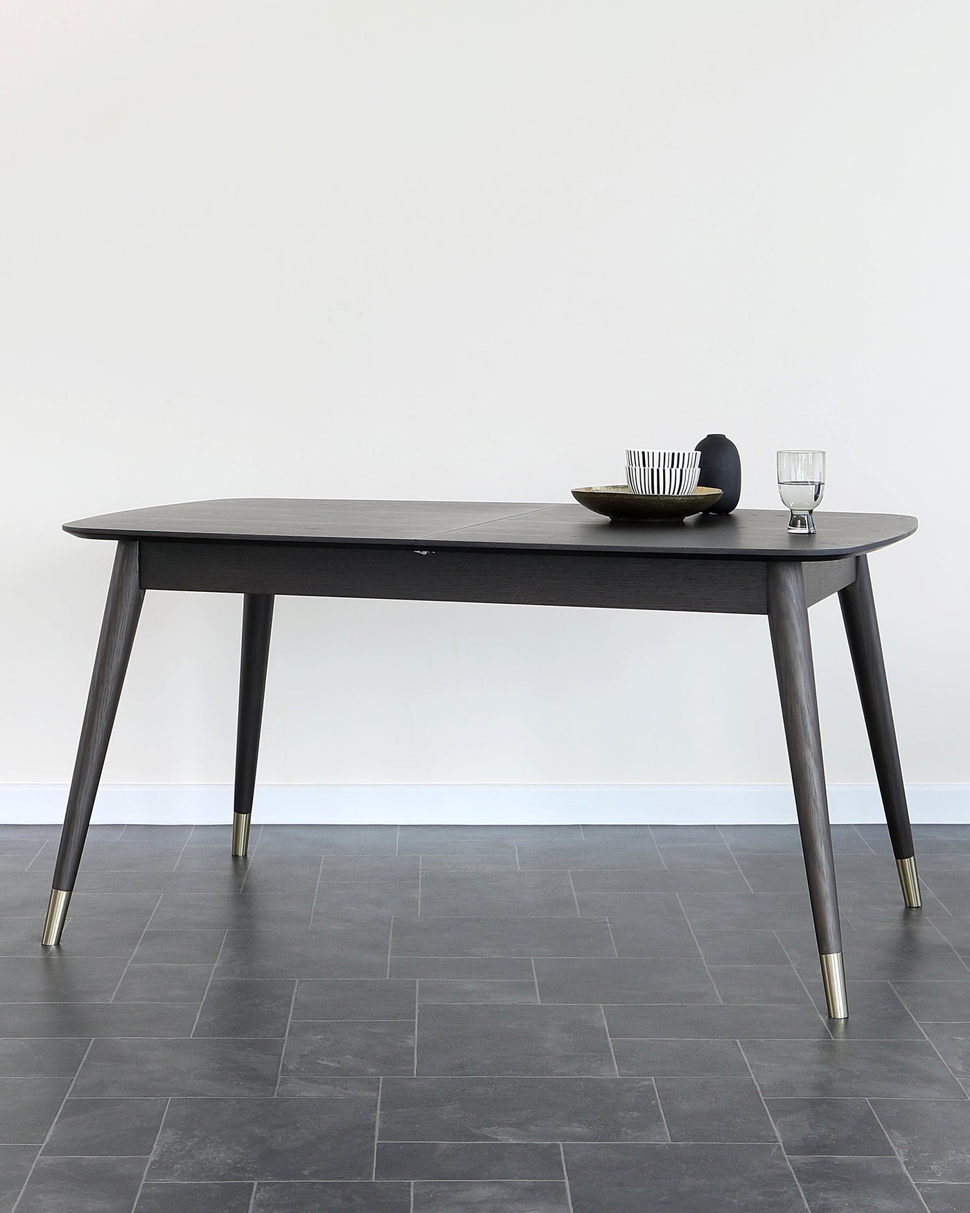 An elegant, mid-century modern style dining table with a dark wood finish and tapered legs, complemented by brass-capped feet. The table, set against a wall with a light grey floor, is adorned with a minimalist centrepiece and a clear glass.