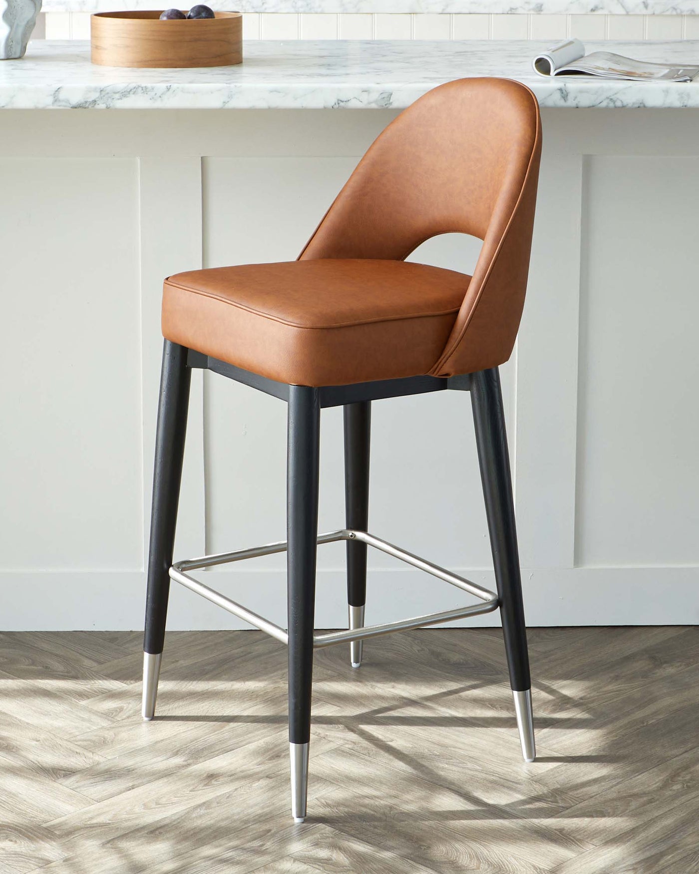 Modern bar stool with a caramel brown upholstered seat and curved backrest, mounted on a black metal frame with sleek metallic accents on the legs and a footrest for added comfort. The stool is positioned on a herringbone wood floor next to a kitchen counter with a marble top.