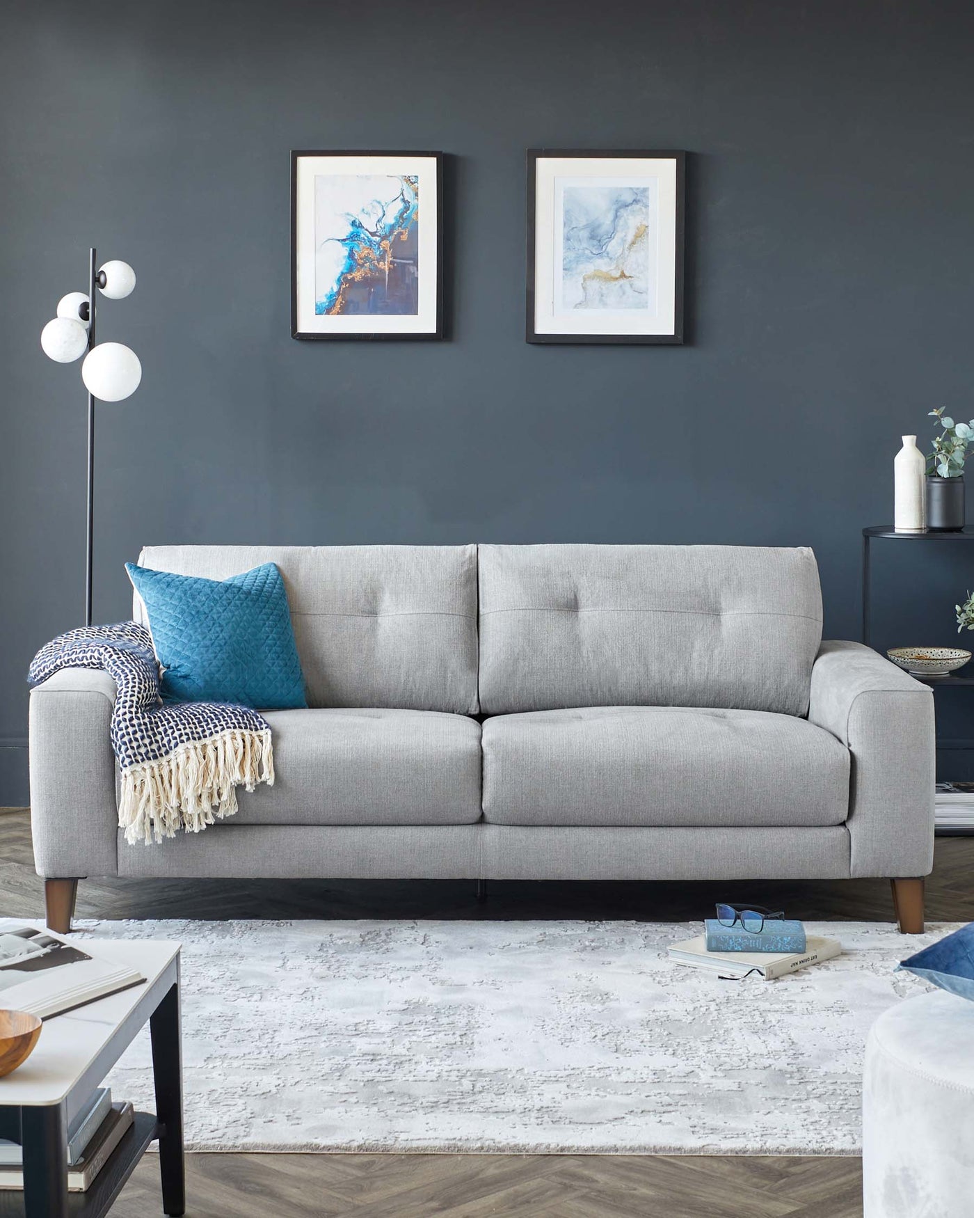 Contemporary grey fabric three-seater sofa with cushioned back and arms, accented with a blue decorative pillow and a patterned white and blue throw blanket. A sleek black floor lamp with a minimalist design featuring globe lights stands to the left, and a dark-toned side table with a vase holds a sprig to the right. The sofa sits on a distressed white area rug over a dark hardwood floor, complemented by a black coffee table with a stack of books and glasses in the foreground.
