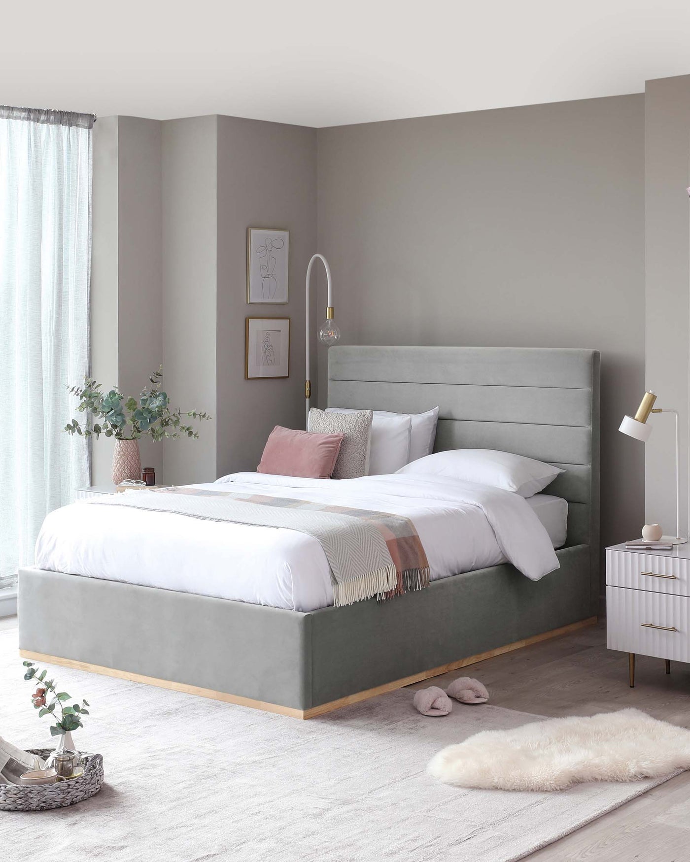 Modern bedroom furniture set featuring a king-sized bed with a tufted, upholstered headboard in a soft grey fabric and a matching platform base with wooden accents. A stylish white bedside table with gold accents and slender legs is placed next to the bed.