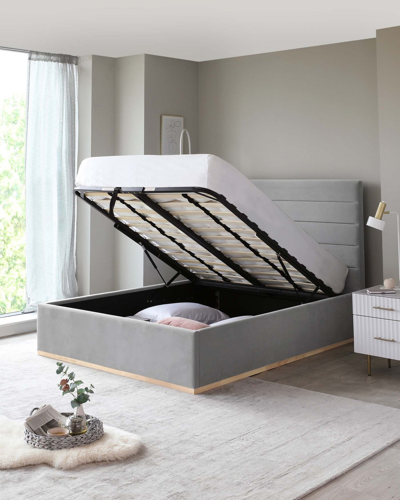 Modern grey upholstered storage bed with a lifted frame revealing under-mattress space on a light wooden floor, accompanied by a sleek white bedside table with golden handles. A cosy rug, soft throw, and decorative items create an inviting atmosphere.