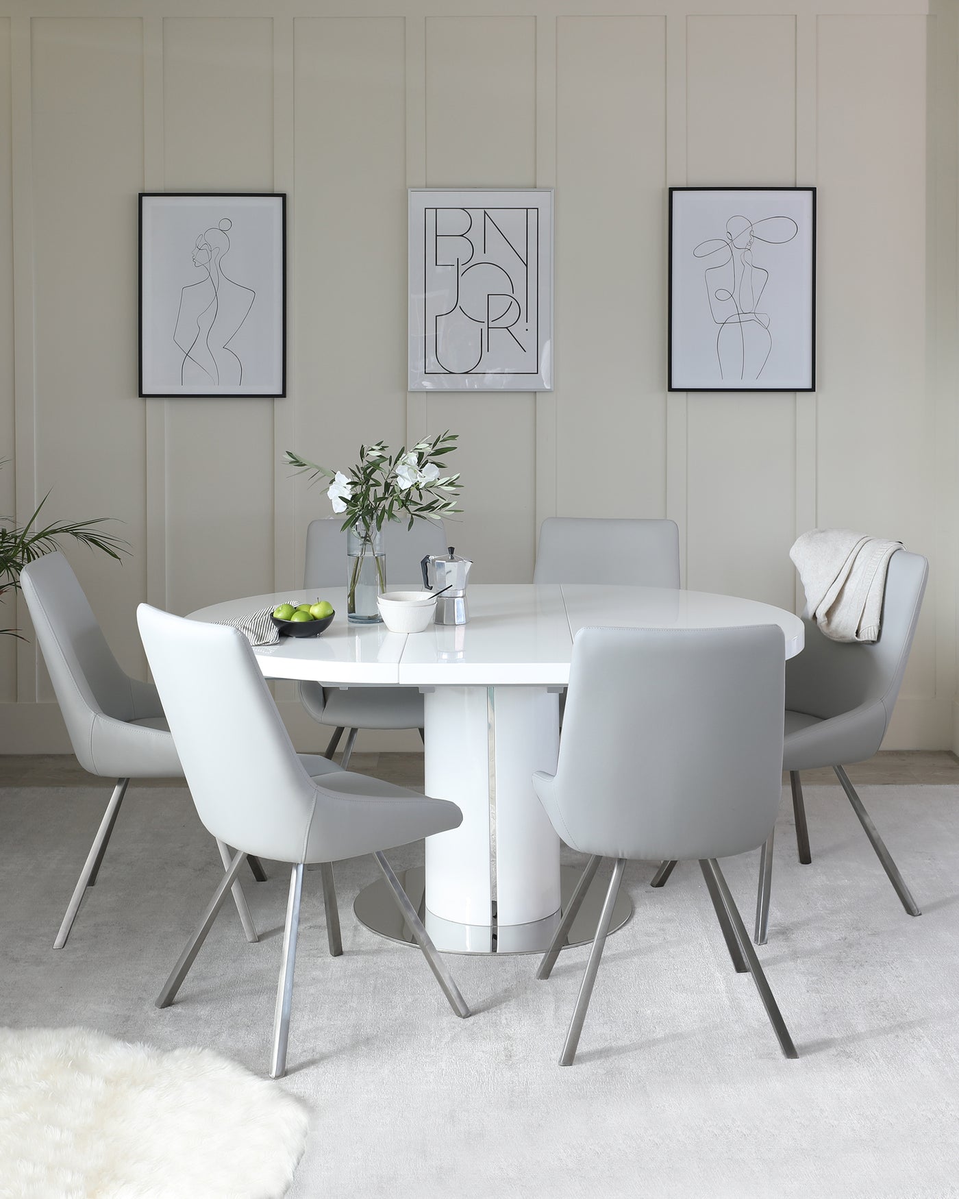 Modern dining room furniture featuring a white, round dining table with a reflective metal base, surrounded by six light grey, sleek upholstered chairs with slender metal legs. A cosy white fur rug lies beneath, adding warmth to the space.